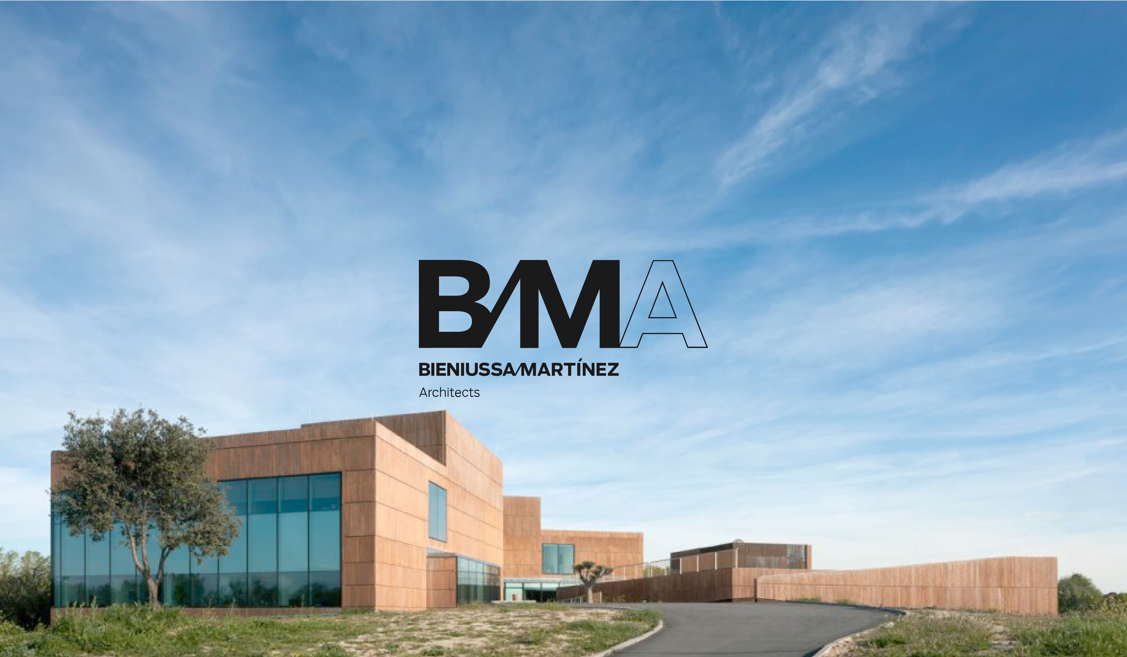 The image shows a modern architectural building under a blue sky with scattered clouds. The structure has large glass windows and an earthy, brown exterior. A curved pathway leads up to the entrance. The logo "BMA Bieniussa-Martínez Architects" is overlaid on the sky.
