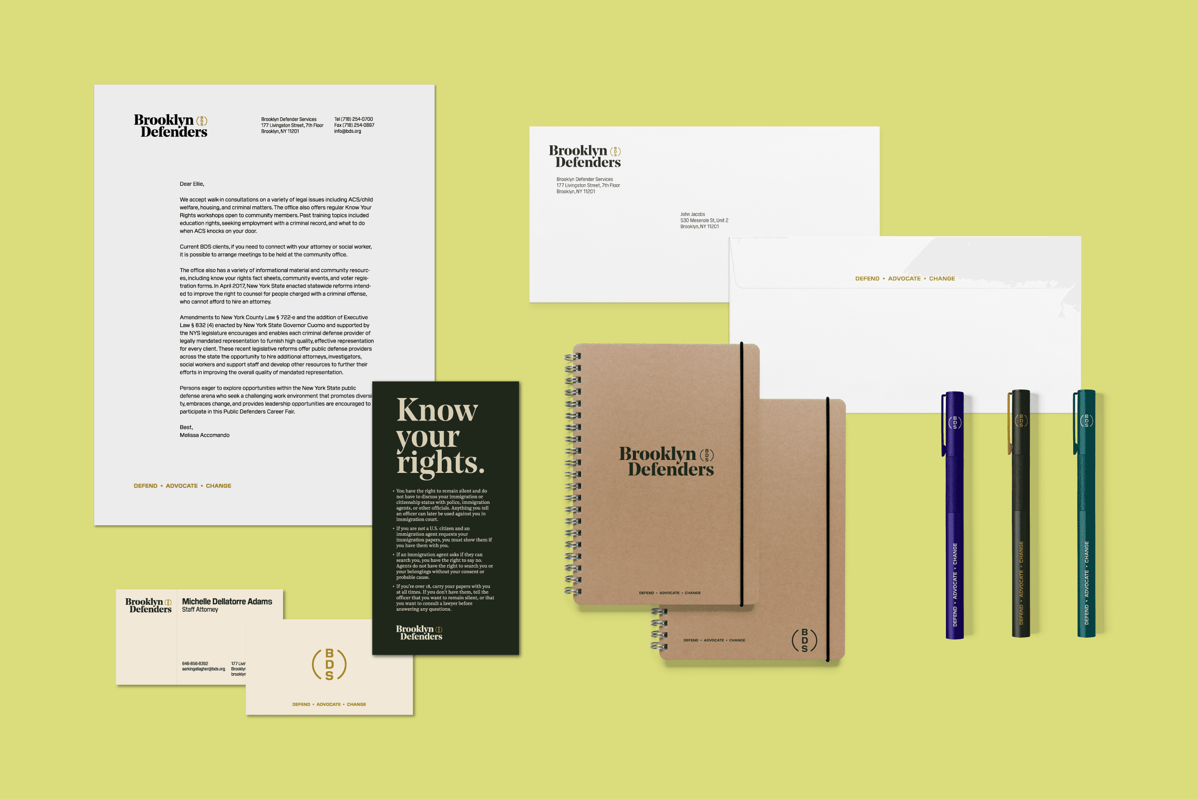 A set of office stationery items is displayed on a light green background, including two spiral-bound notebooks, three pens in blue, black, and green, a letterhead, business cards, an envelope, and informational cards. The items are branded with "Brooklyn Defenders.
