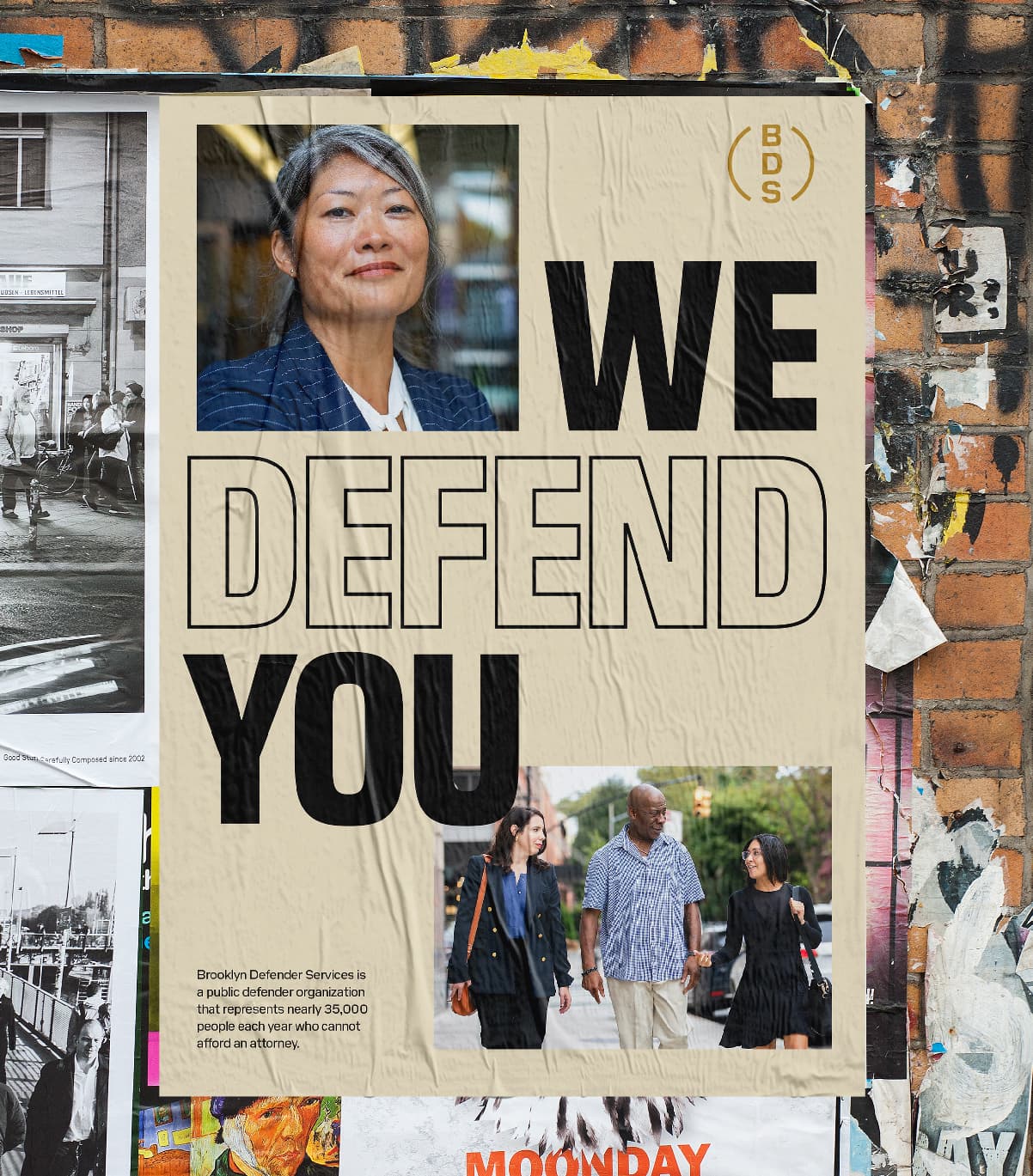 A wall poster displays a smiling woman above the large text "WE DEFEND YOU." Below the text, there is an image showing three people walking together. The poster is surrounded by other overlapping posters, with a brick wall background.