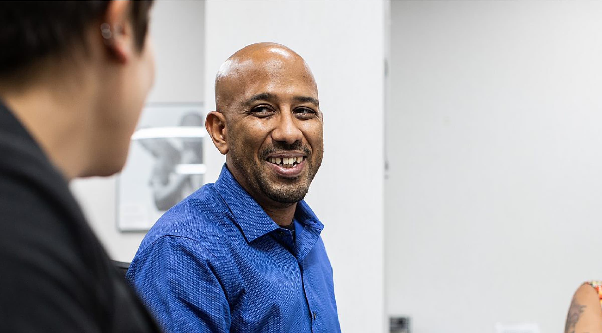 A man wearing a blue shirt smiles during a conversation with a colleague. They are indoors, seated in an office setting with light-colored walls and a picture frame in the background. Only the side profile of the colleague is visible.