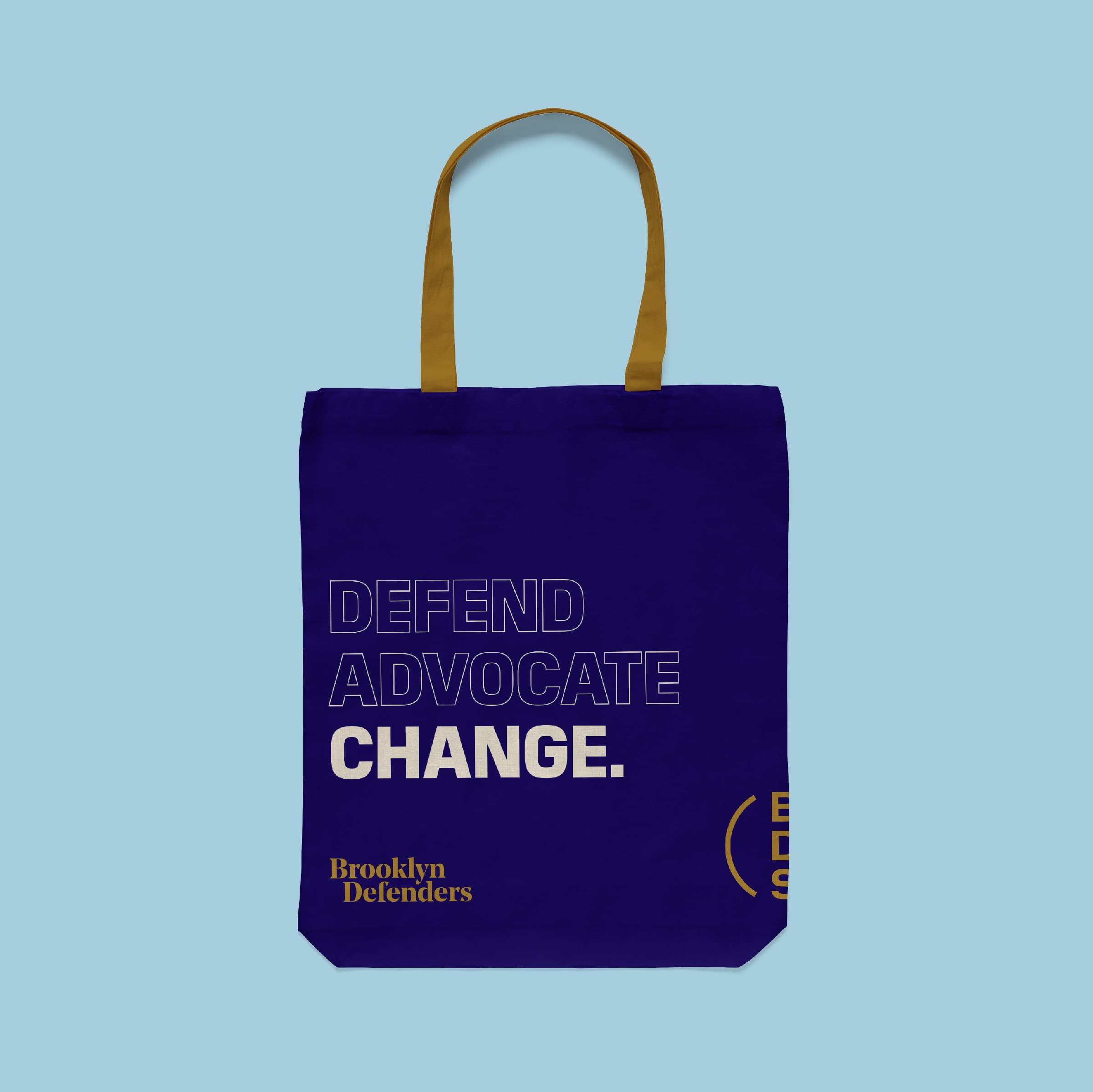 A blue tote bag with brown handles is pictured against a light blue background. The bag features the words "DEFEND ADVOCATE CHANGE" in large white letters, with "Brooklyn Defenders" printed in yellow text below.