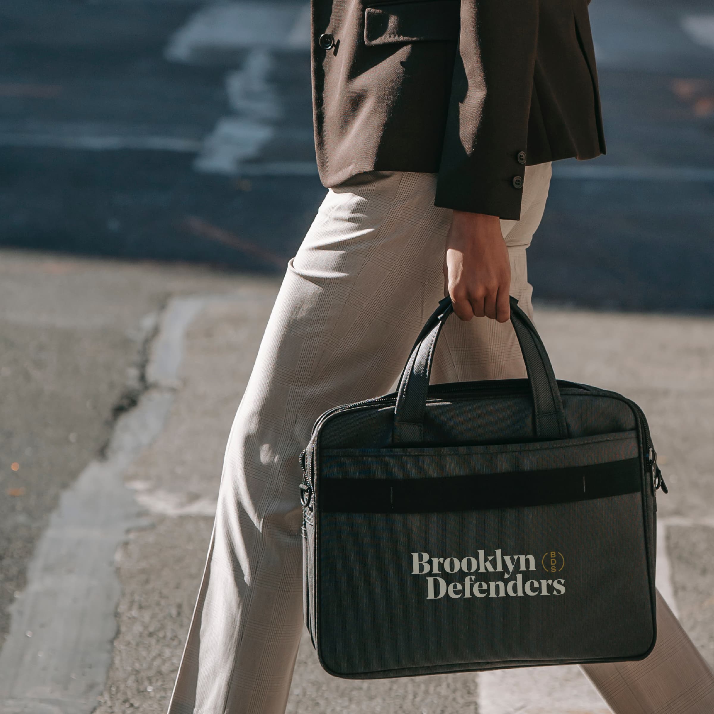 A person dressed in business attire is walking down a street carrying a black briefcase with the words "Brooklyn Defenders" printed on it. The briefcase has two short handles and appears to be made of fabric. The background features a crosswalk and paved road.