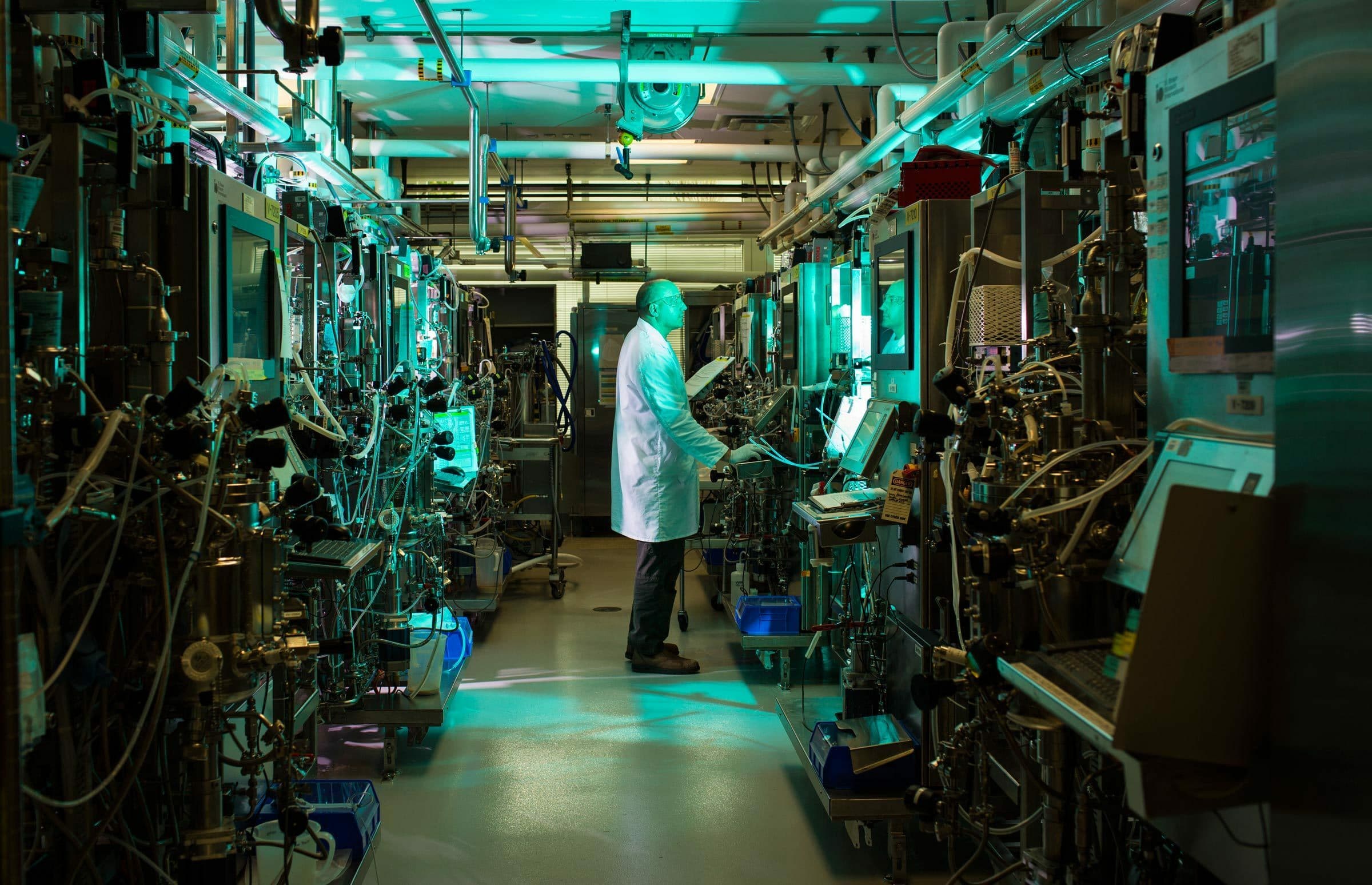 A scientist wearing a lab coat stands in a narrow, dimly lit lab filled with large machines and complex equipment, all casting an eerie green light. He interacts with a screen, appearing focused amidst the intricate array of pipes, wires, and control panels.