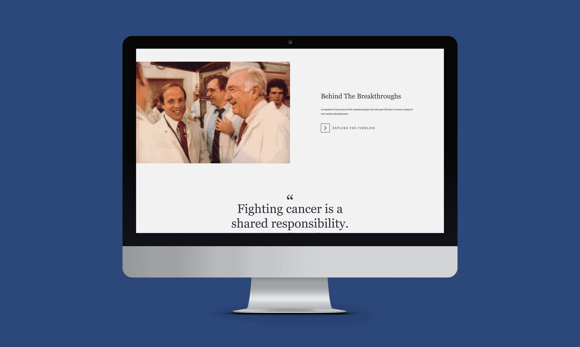 A computer monitor displays a webpage featuring an image of several people in lab coats smiling and interacting. The text on the screen says, "Behind The Breakthroughs" and "Fighting cancer is a shared responsibility." There is a button labeled "Explore the Timeline.
