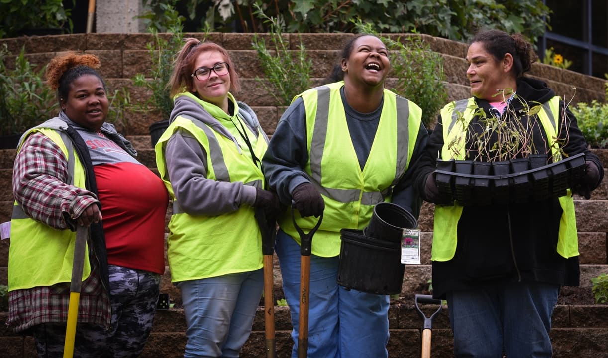 Four people dressed in yellow safety vests are standing in a garden, smiling and holding gardening tools and plants. A tiered stone wall with greenery is in the background. They appear to be enjoying their time participating in an outdoor gardening activity.