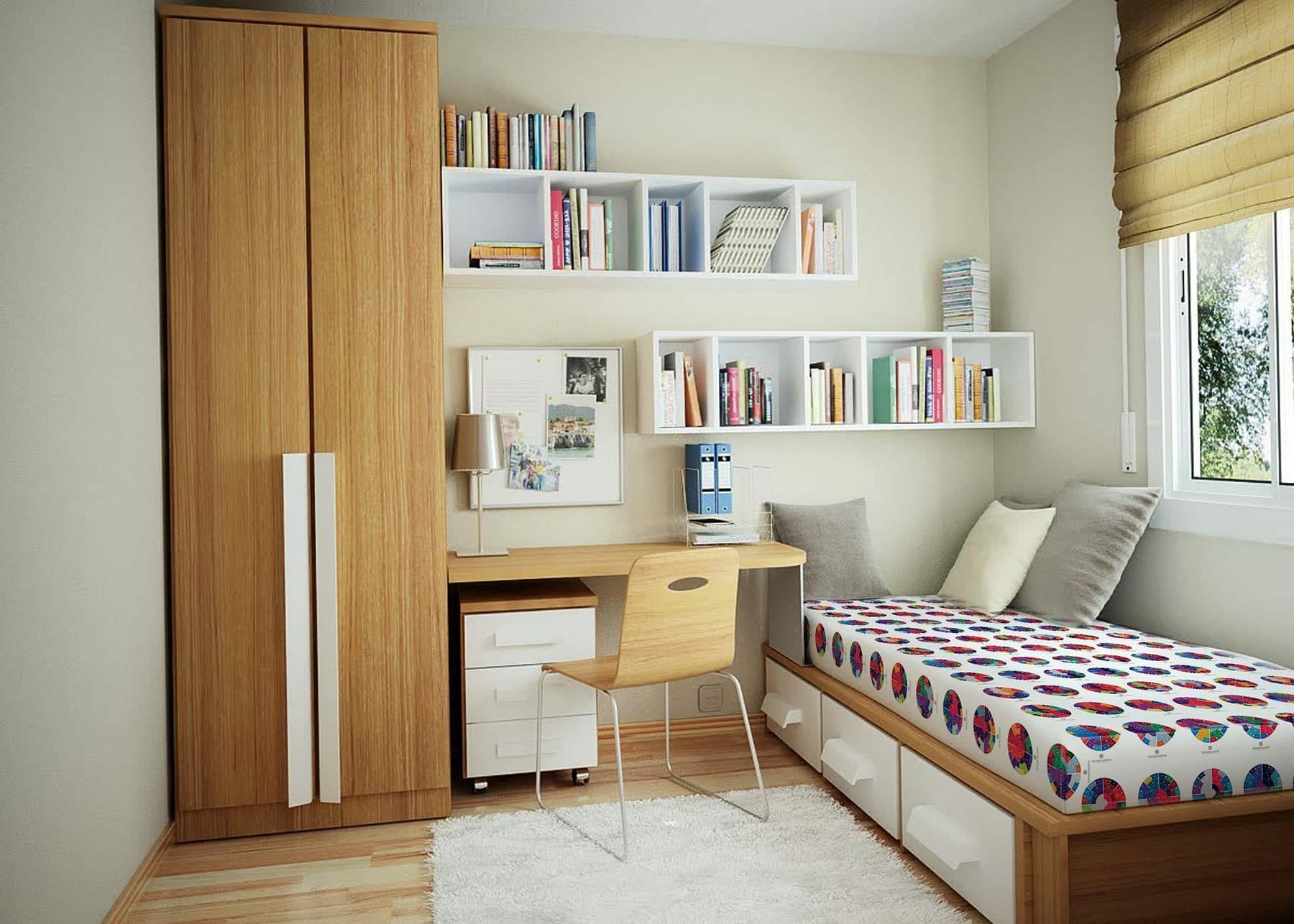 A small, cozy bedroom with light-colored walls features a wooden wardrobe, a white and wooden desk with books and a pinboard, and wall-mounted white shelves filled with books. A single bed with a colorful patterned duvet, drawers underneath, and a window with beige blinds completes the room.