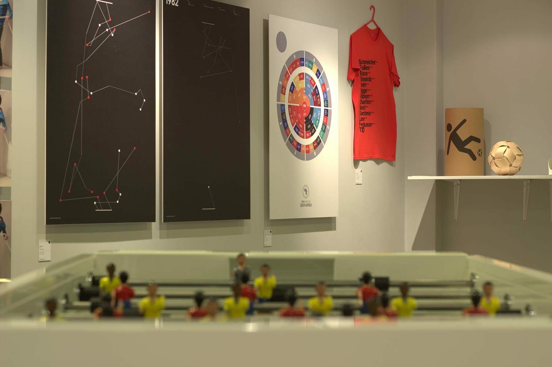 A photography exhibit features abstract art pieces on the wall, a red t-shirt on a hanger with text, a white shelf displaying a vinyl record case and a soccer ball. In the foreground, a foosball table with colorful players is partially visible.