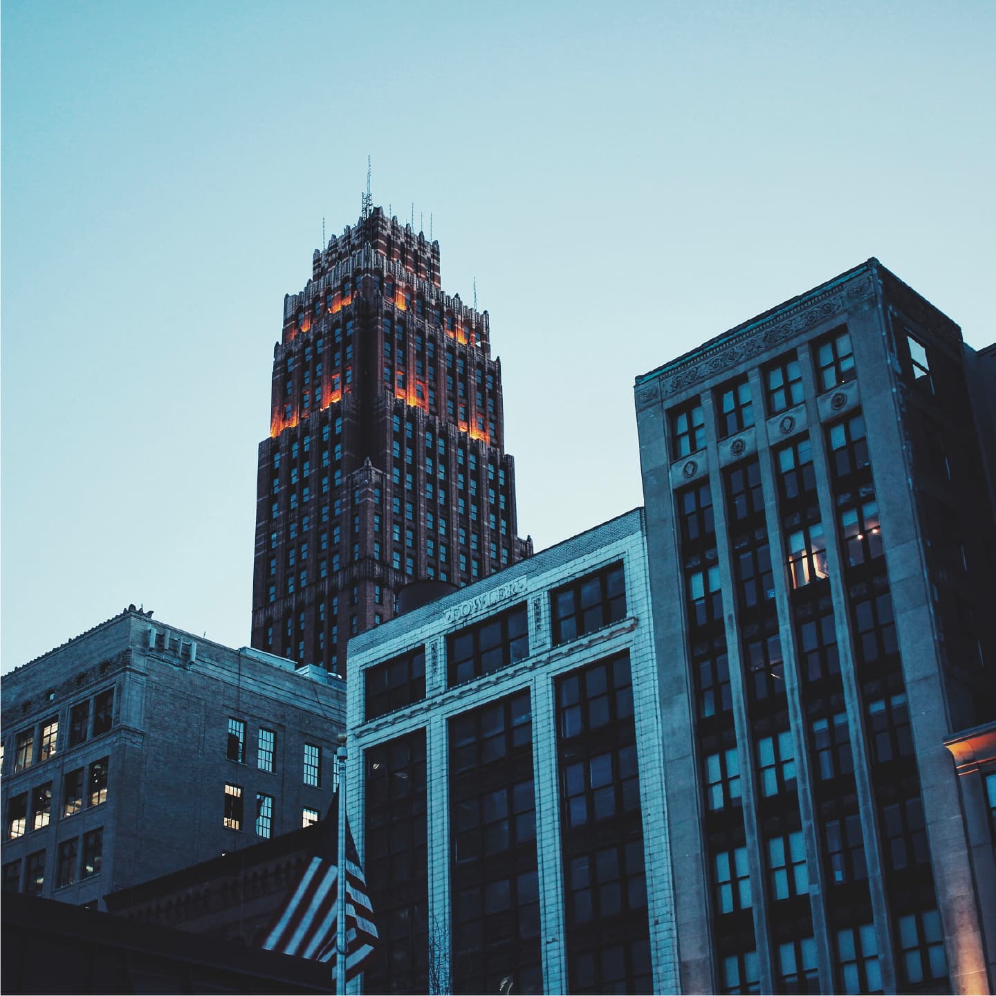 A tall, illuminated skyscraper stands in the background with a dimly lit facade, surrounded by shorter buildings with a mix of modern and classic architectural styles. An American flag is visible in the lower left corner. The sky is clear and light blue.