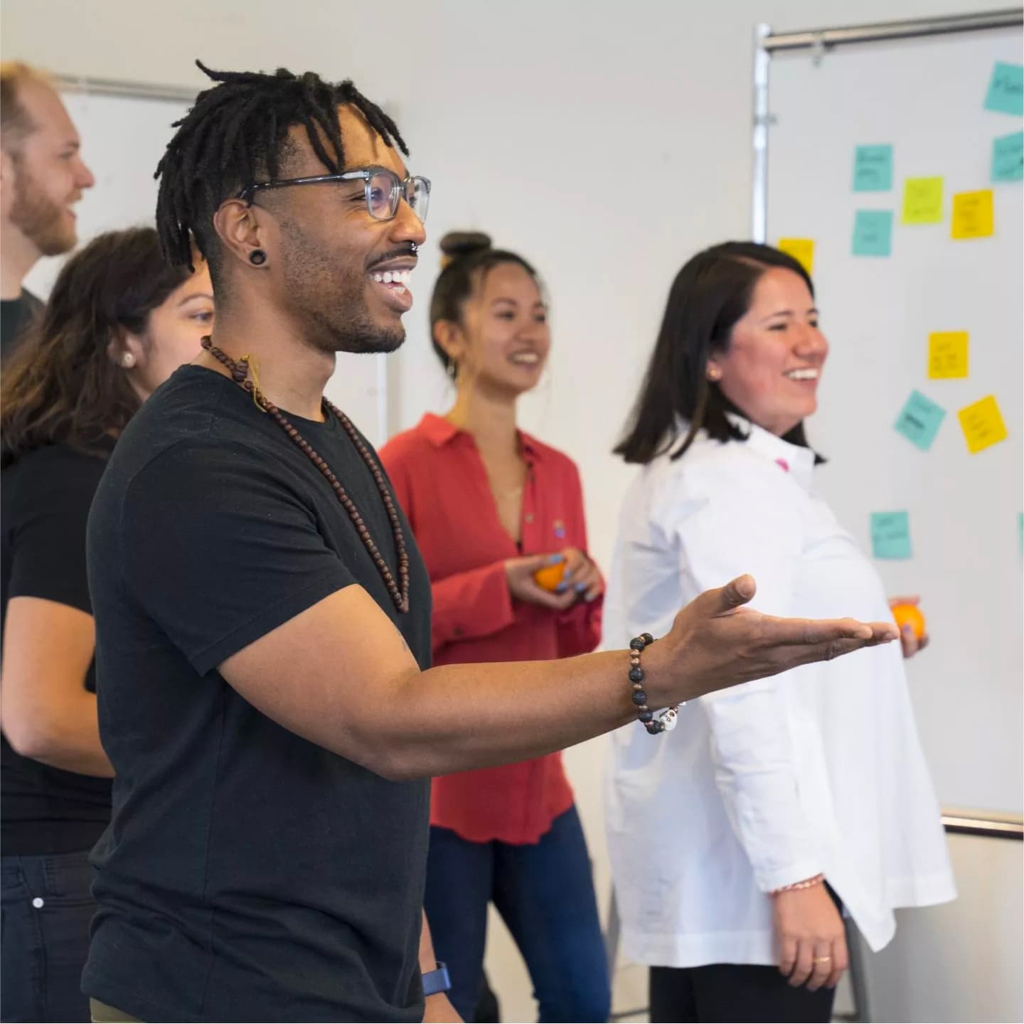 A diverse group of five people are smiling and engaging in a collaborative activity in an office. One individual in the foreground is gesturing with his hand. Behind them is a whiteboard covered with colorful sticky notes.