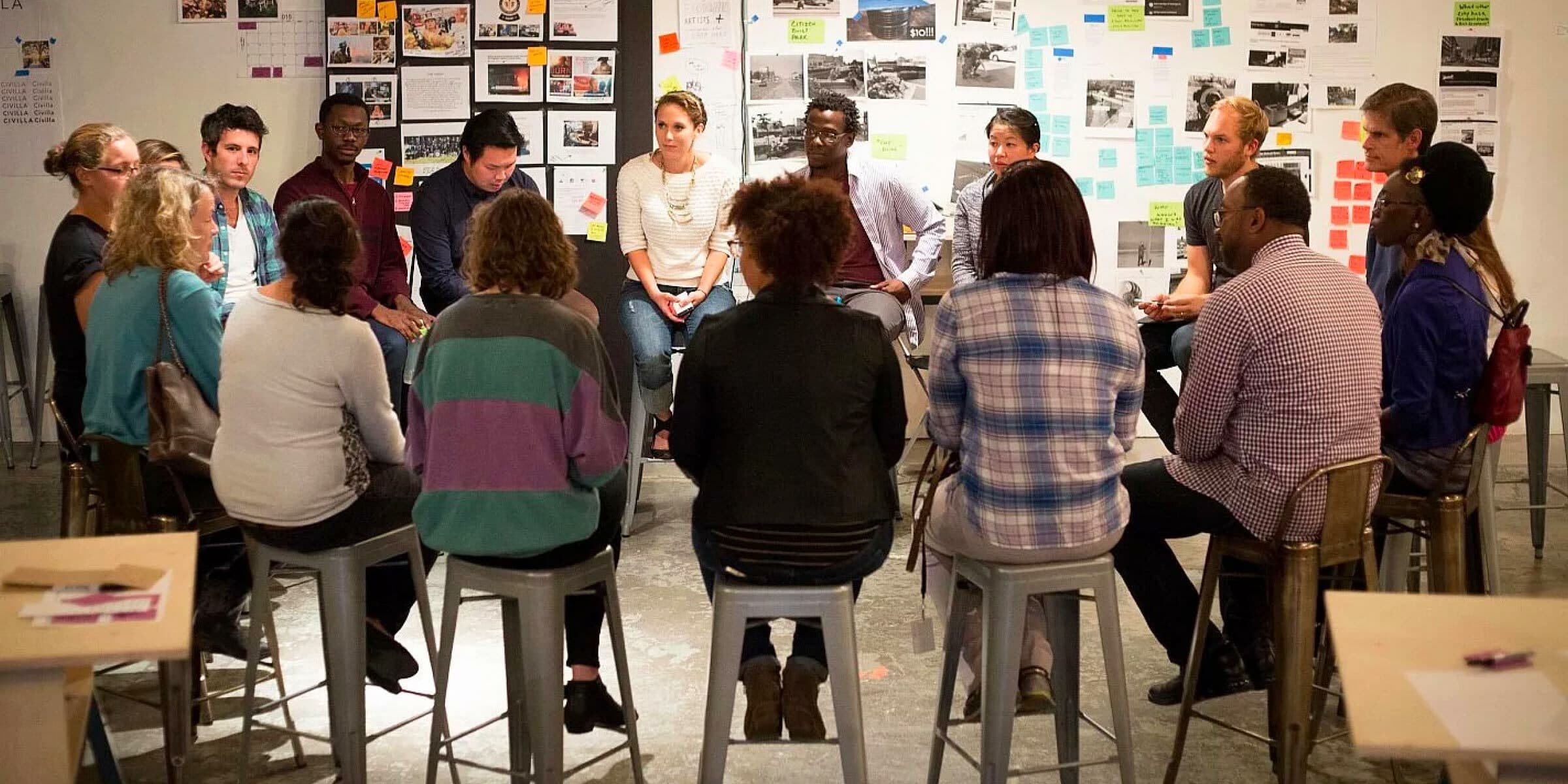 A diverse group of people sits in a circle on stools in a brightly lit room, engaged in a discussion. The walls are covered with notes, posters, and images. The atmosphere appears collaborative and focused.