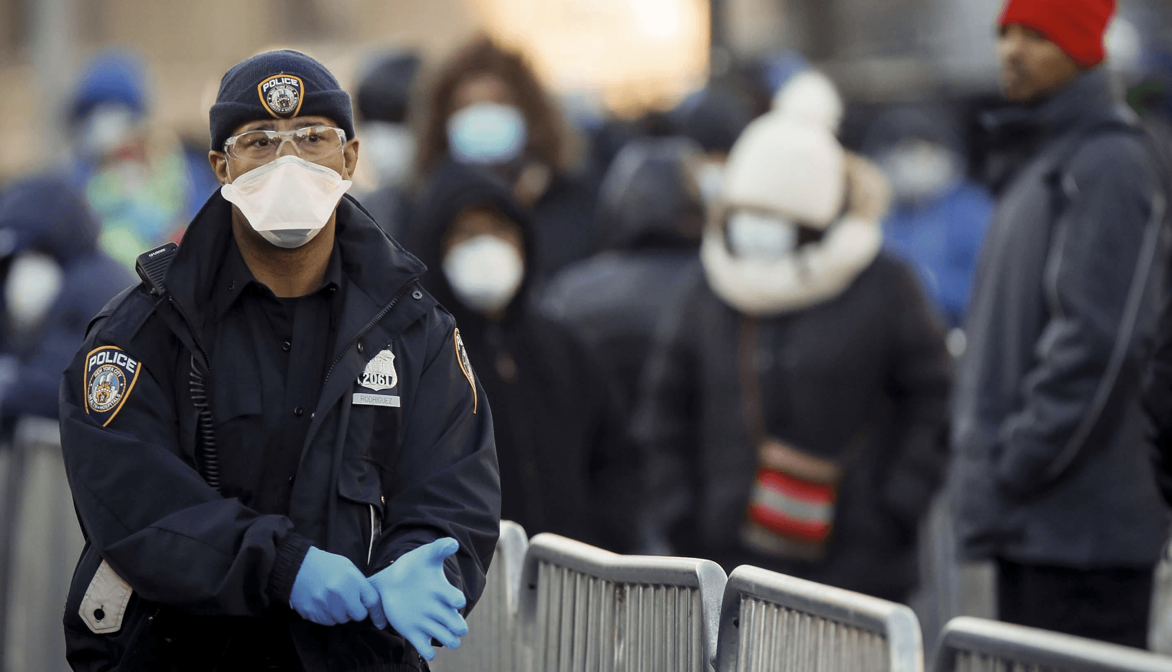 A police officer wearing a face mask, protective eyewear, a beanie, and blue gloves stands in front of metal barriers. In the background, a blurred crowd of people, also wearing masks and winter clothing, are gathered.