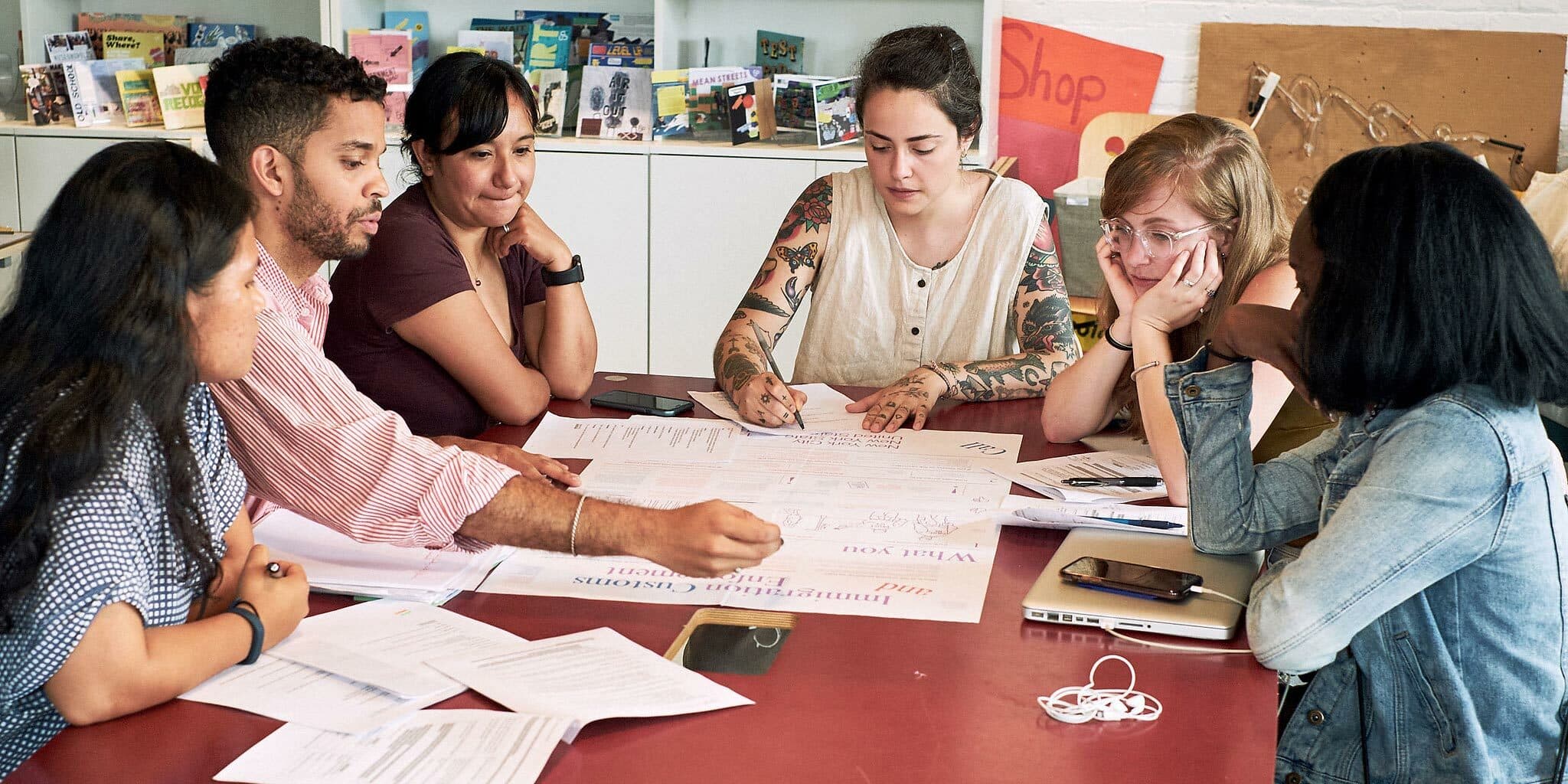 A group of six people sit around a table covered with papers and documents. They are engaged in discussion, with one man pointing at a document. Books and shelves are visible in the background. The atmosphere appears collaborative and focused.