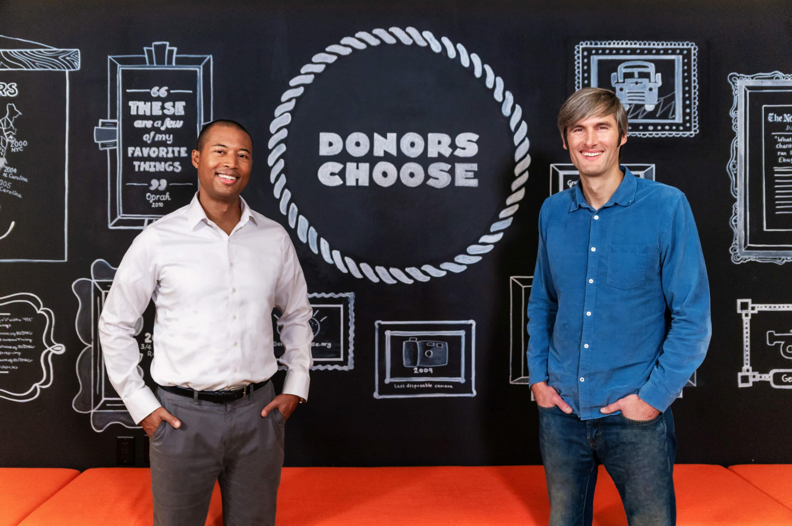 Two people standing in front of a blackboard wall with white chalk drawings and the words "Donors Choose" in the center. The person on the left is wearing a white shirt and gray pants, and the person on the right is wearing a blue shirt and jeans. They are both smiling.
