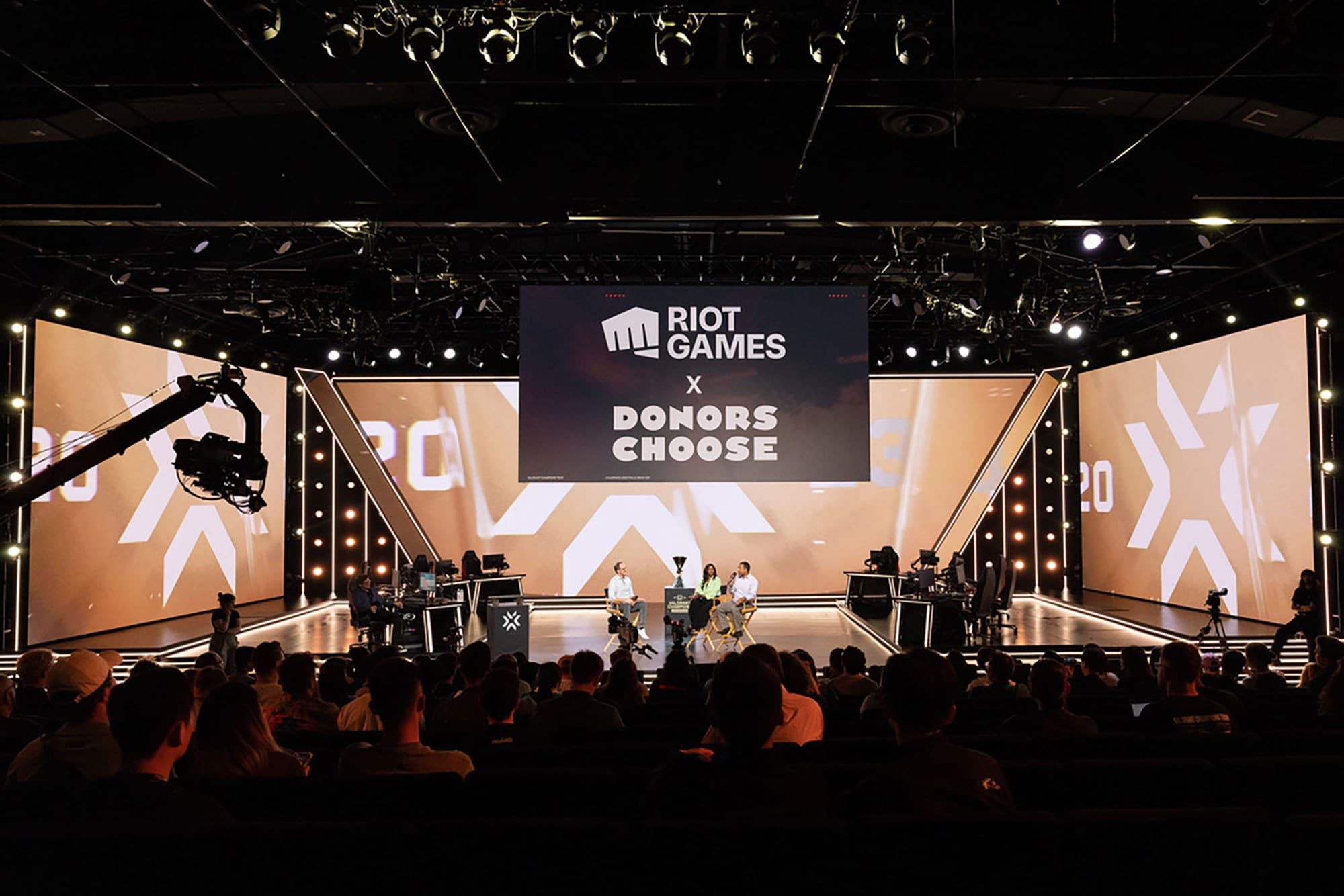 A large audience sits in a modern auditorium facing a well-lit stage with a big screen displaying "Riot Games x Donors Choose." Several people are seated on stage, and cameras are set up for filming.