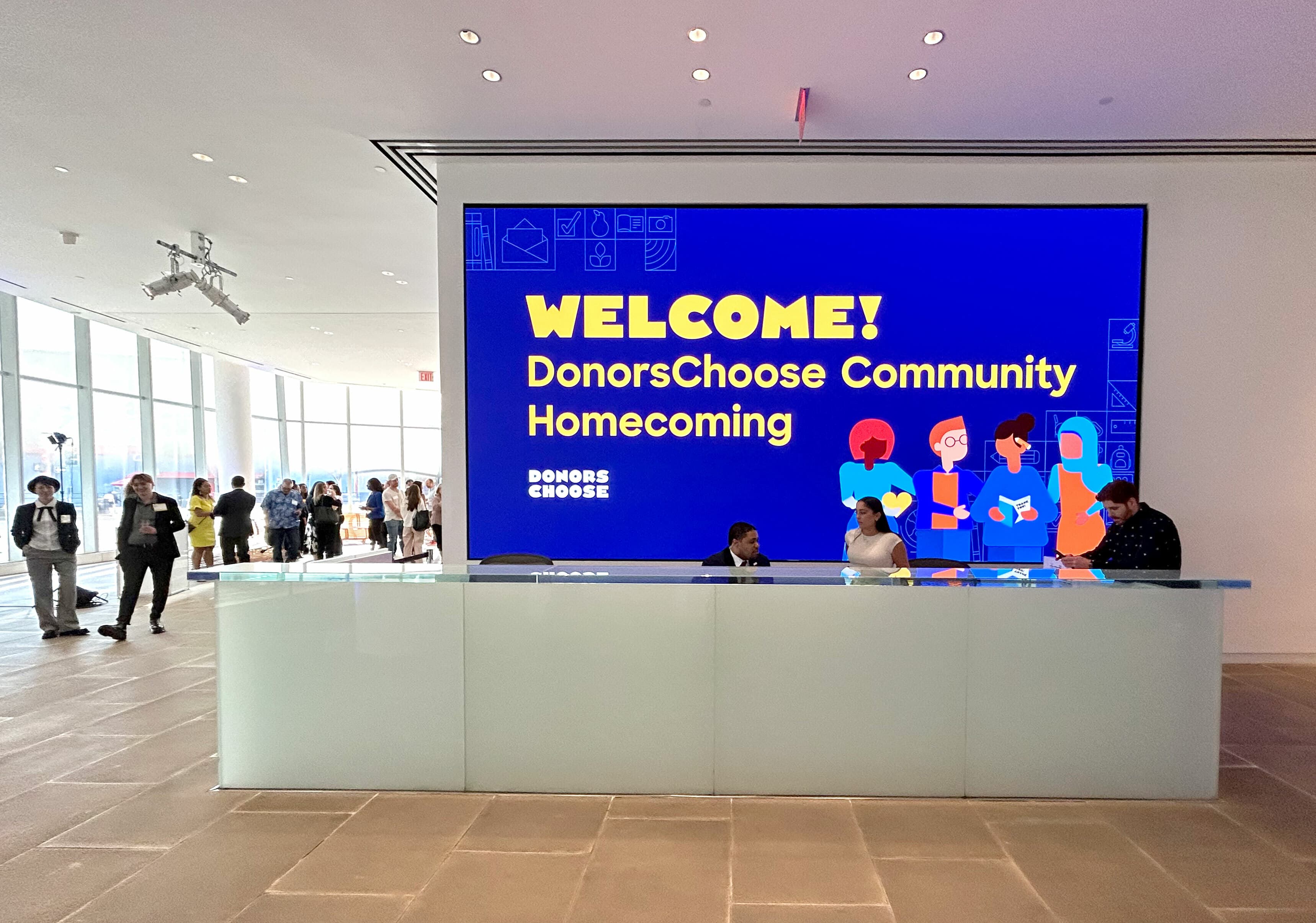 A brightly lit lobby features a large digital screen displaying "WELCOME! DonorsChoose Community Homecoming" with colorful illustrations of diverse individuals. In the foreground, a glass reception desk is staffed by three people. Several attendees stand in the background.