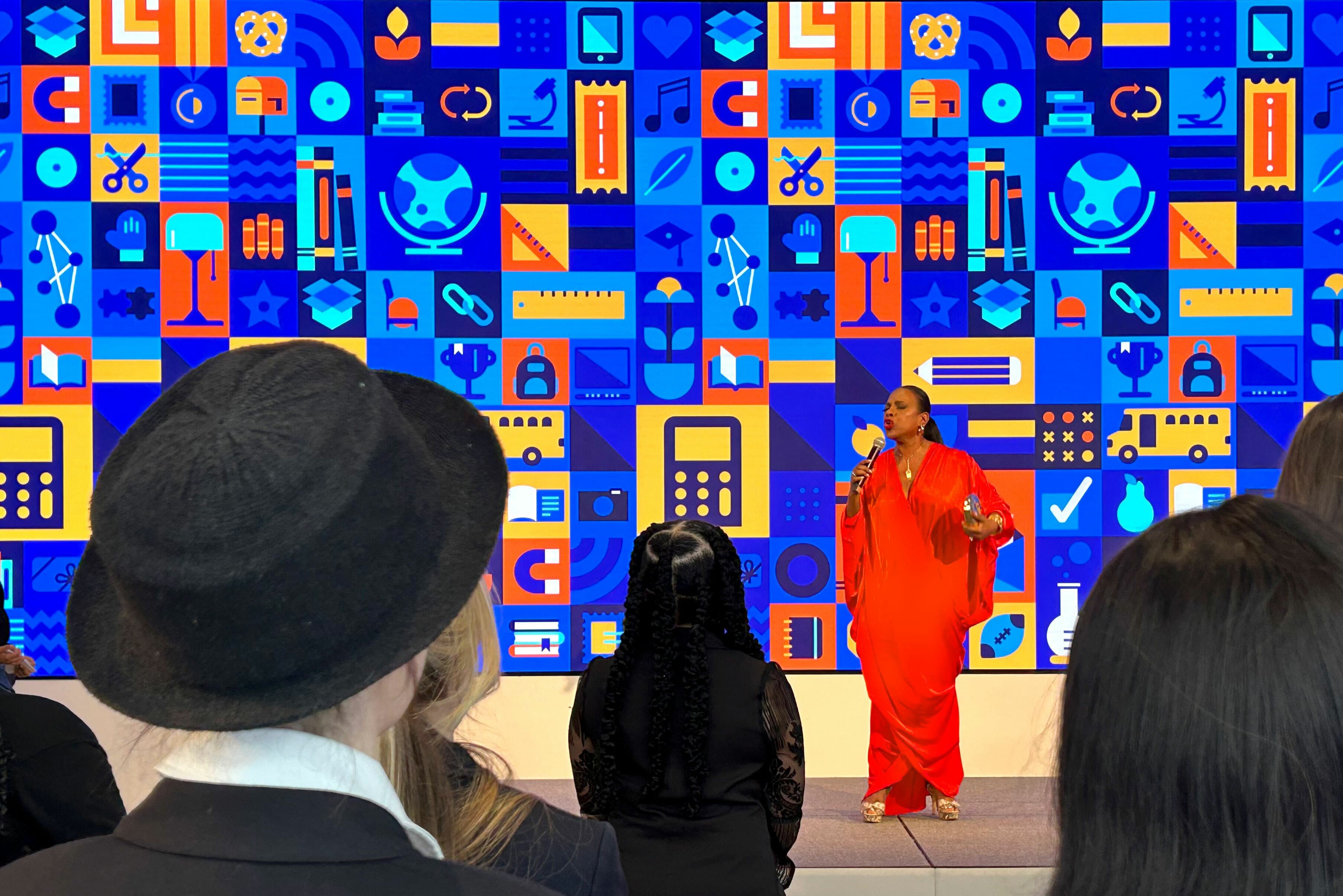 A person in a vibrant red outfit speaks into a microphone on stage in front of a colorful, geometric backdrop. The audience listens attentively, with some members wearing hats and dark clothing. The backdrop features various icons in blue, orange, and yellow hues.