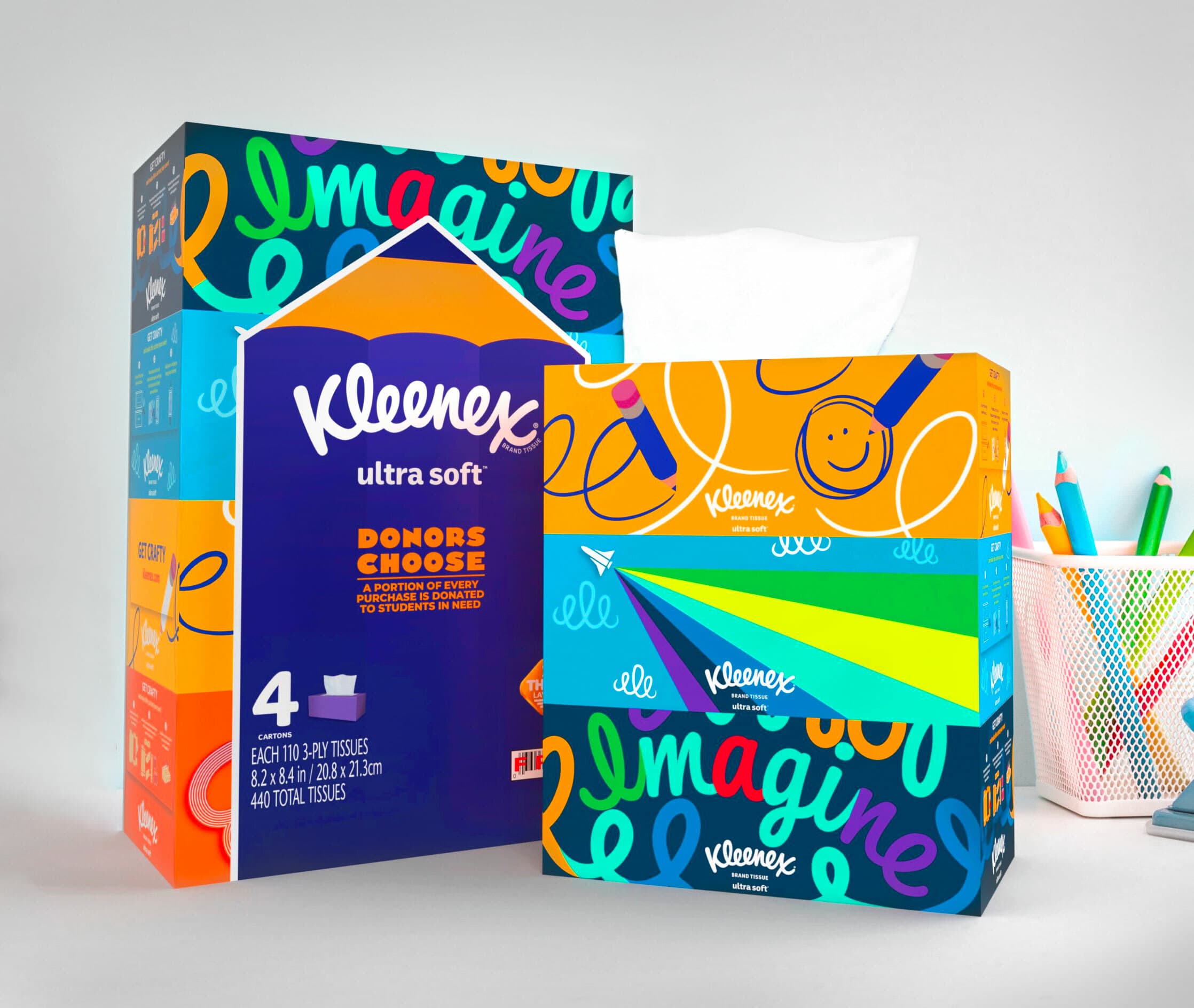 A box and a packaging of Kleenex ultra soft tissues are shown. The packaging features colorful, playful artwork with the word "Imagine." Beside the tissues is a cup holding assorted pens and pencils on a white surface.