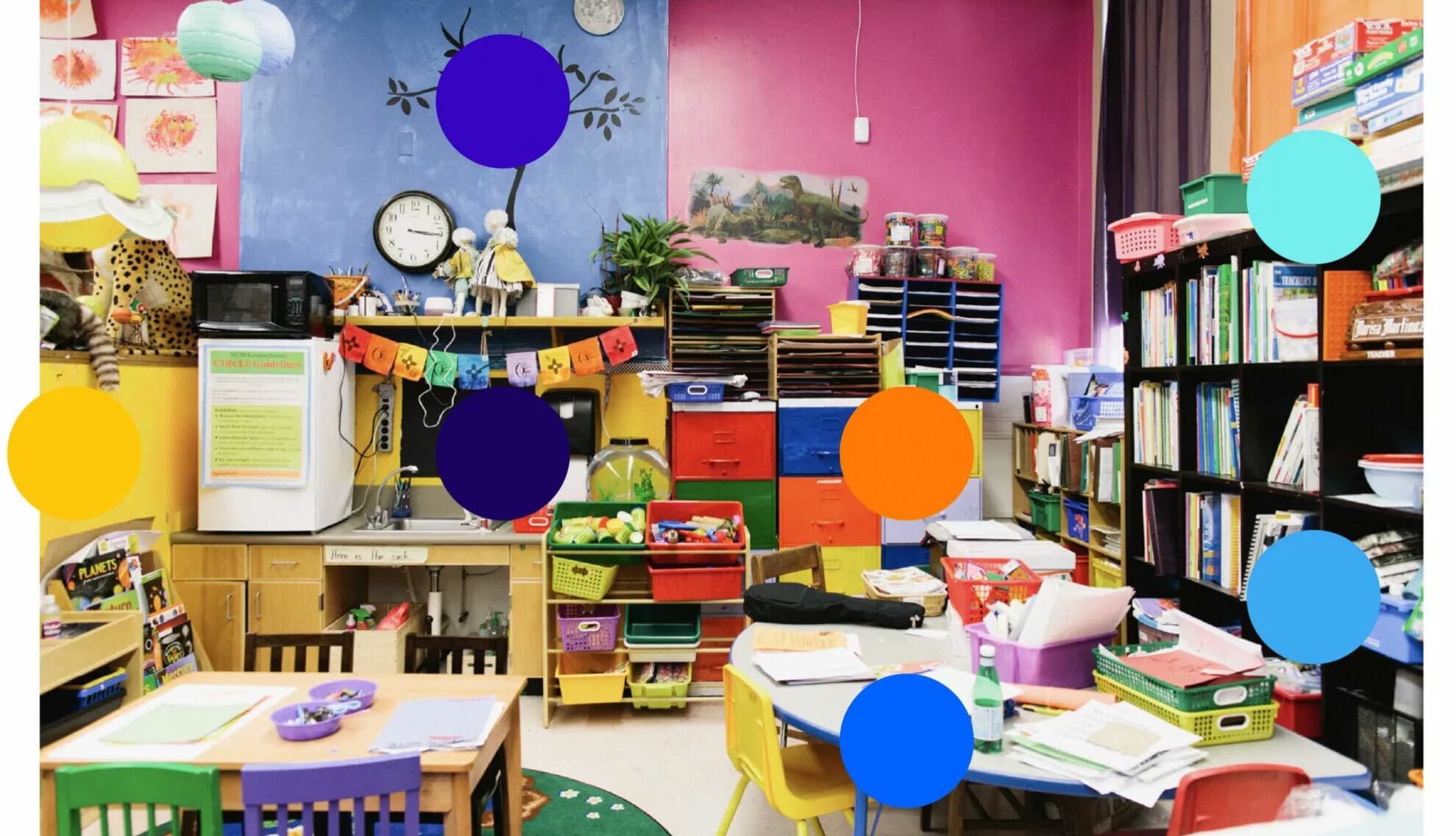 Colorful classroom decorated with vibrant artwork, various storage bins, shelves filled with books, and educational materials. Tables and chairs are arranged for student activities. Bright walls with posters and a potted plant add to the lively environment.