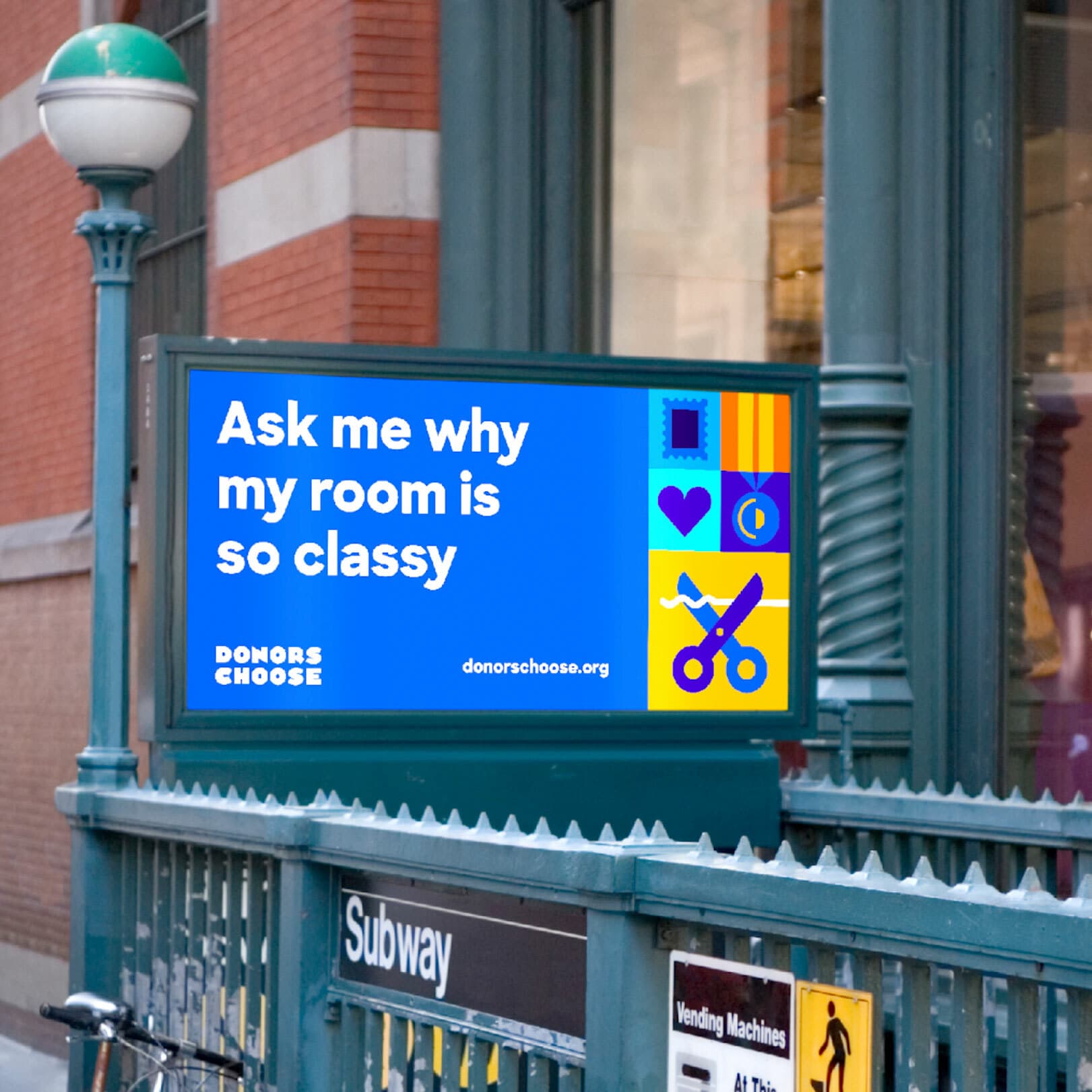 A lit billboard at a subway entrance showcases a DonorsChoose advertisement. It reads, “Ask me why my room is so classy” and features colorful icons, including a heart, book, and scissors, against a blue background. The DonorsChoose website address is also displayed.