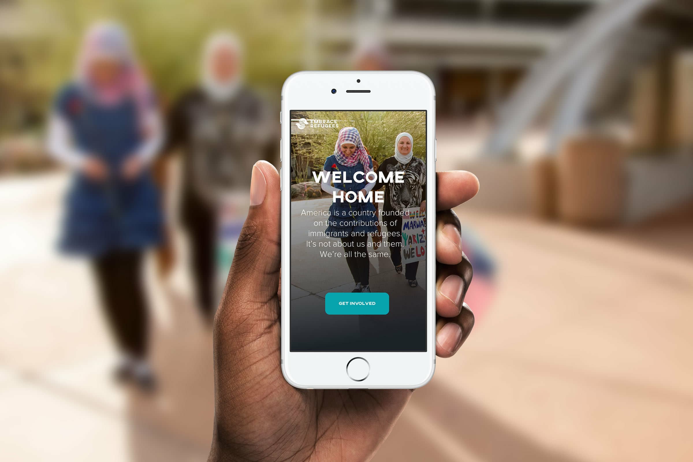 A hand holding a smartphone displaying a website with the text "WELCOME HOME" and "America is a country founded on the contributions of immigrants and refugees. It's not about us or them. We're all the same." The background shows three people walking and talking.