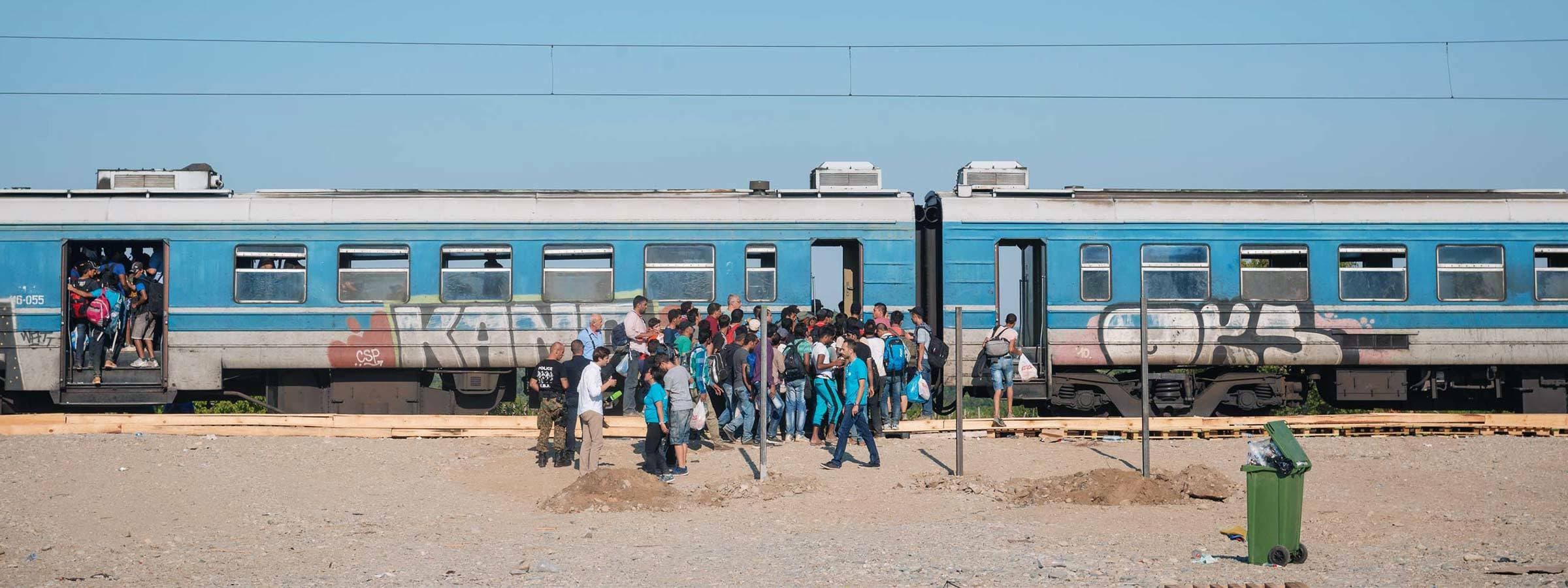 A large group of people are boarding a blue train at an outdoor train station. The train is covered in graffiti, and there is a green trash bin in the foreground. The sky is clear and blue.