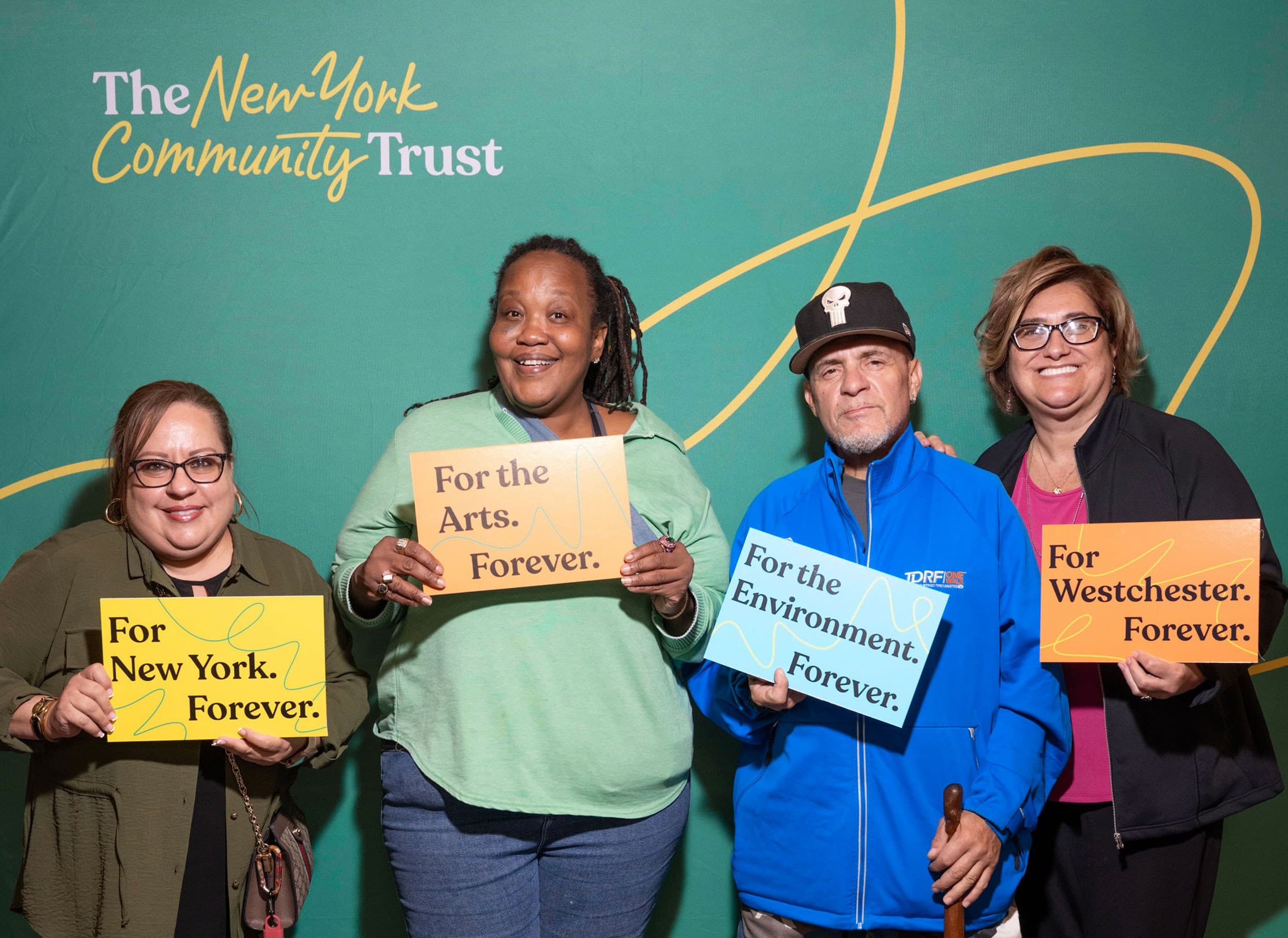 Four people stand smiling in front of a green backdrop with “The New York Community Trust” logo, holding signs that say: “For New York. Forever.”, “For the Arts. Forever.”, “For the Environment. Forever.”, and “For Westchester. Forever.”.