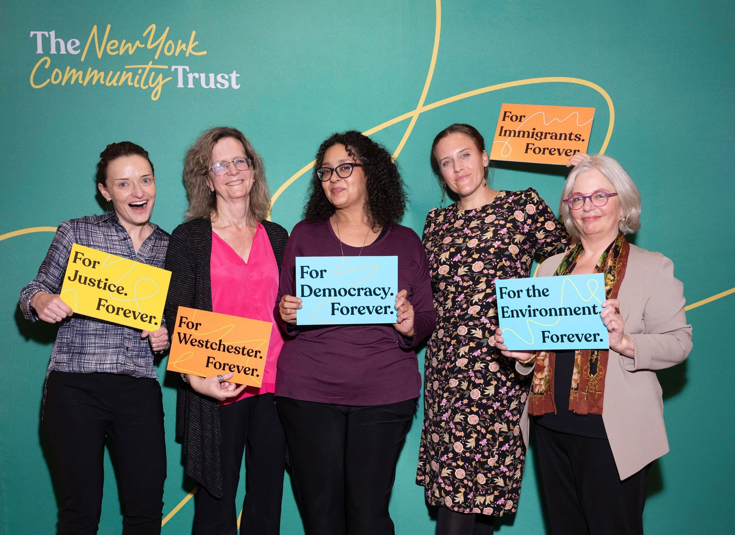 Five women stand in front of a green backdrop with "The New York Community Trust" logo, each holding signs with messages supporting justice, Westchester, democracy, immigrants, and the environment.
