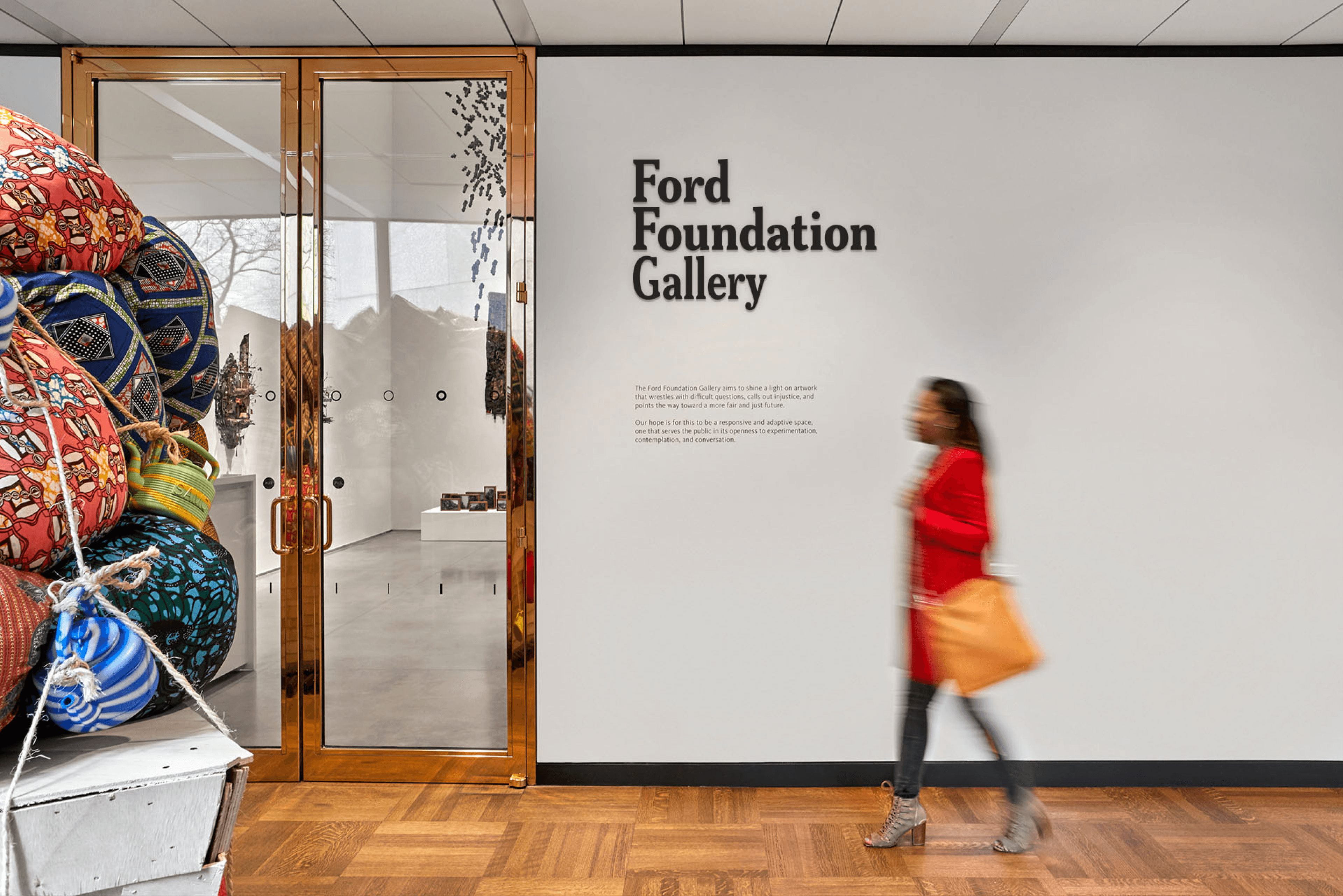 A person wearing a red coat walks past the entrance to the Ford Foundation Gallery. The entrance features glass doors flanked by an art installation of colorful, patterned bundles. The gallery's name and description are displayed on the white wall next to the door.