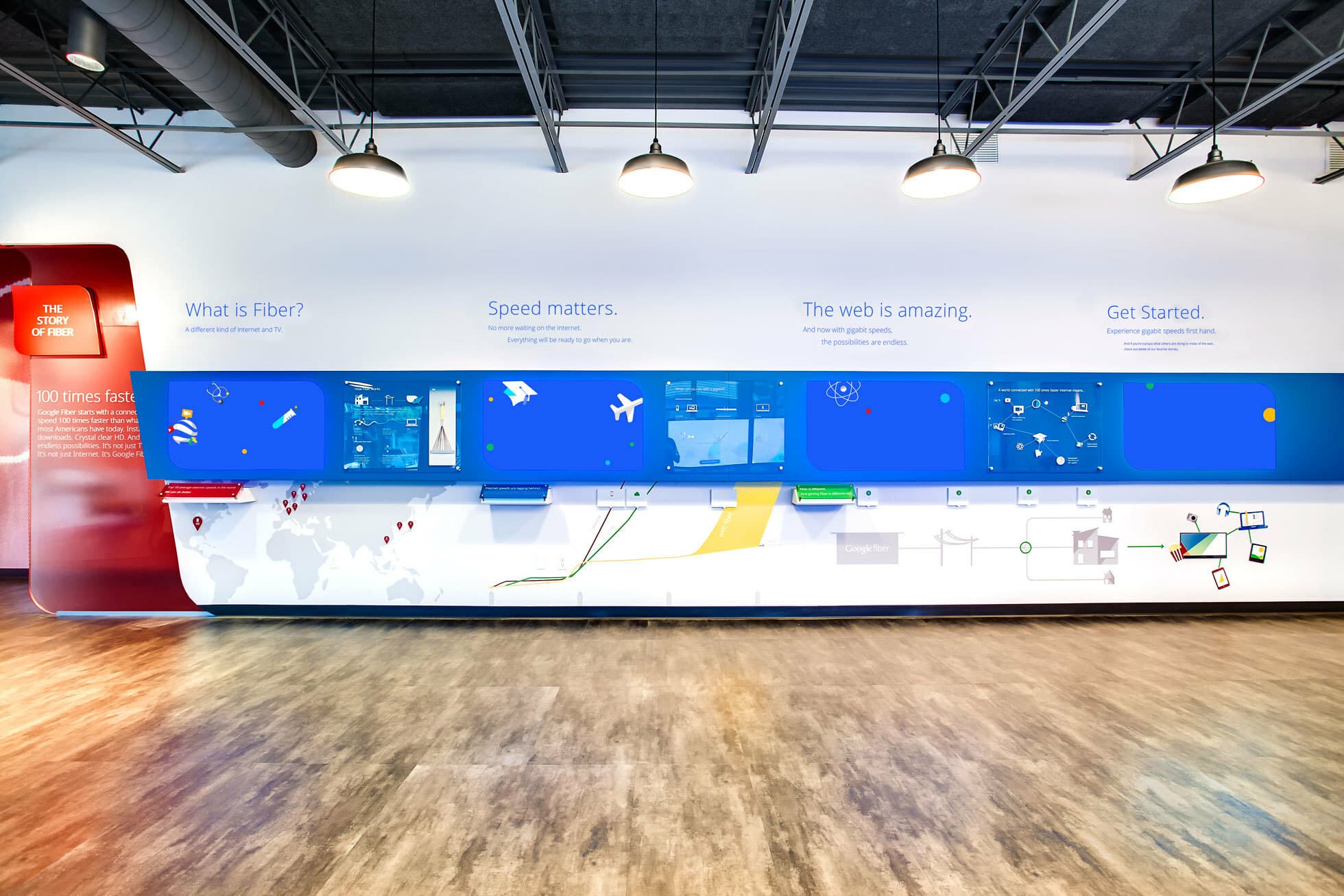 A modern, brightly lit tech display wall features sections on "What is Fiber?", "Speed matters.", "The web is amazing.", and "Get Started." Icons and graphics illustrate concepts, with various interactive elements and maps below. Ceiling lights illuminate the space.