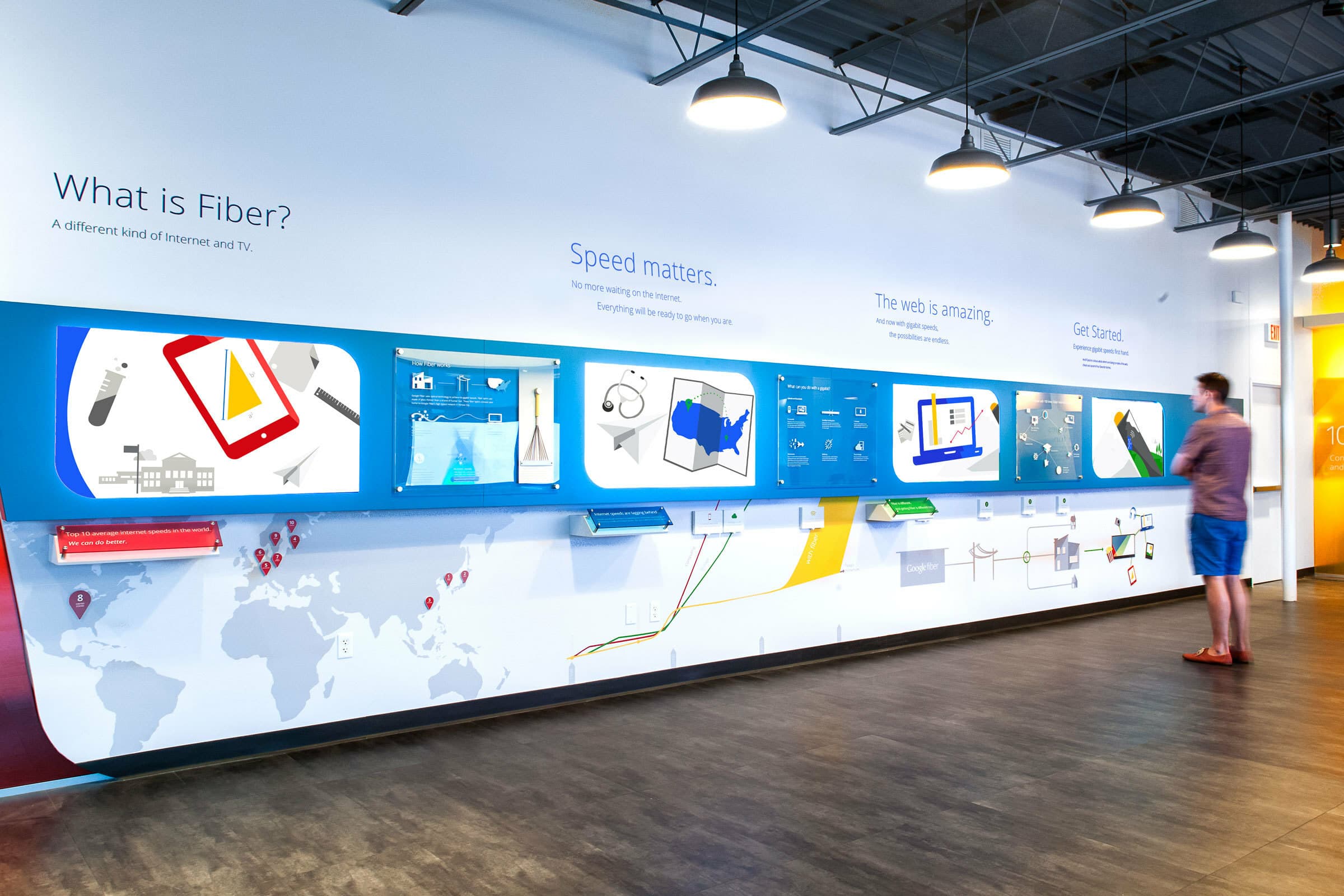 A person stands in front of an interactive wall display explaining fiber optics. The wall showcases colorful diagrams and maps about internet speed and connectivity. The area is lit with modern industrial overhead lighting, and the floor is wooden.