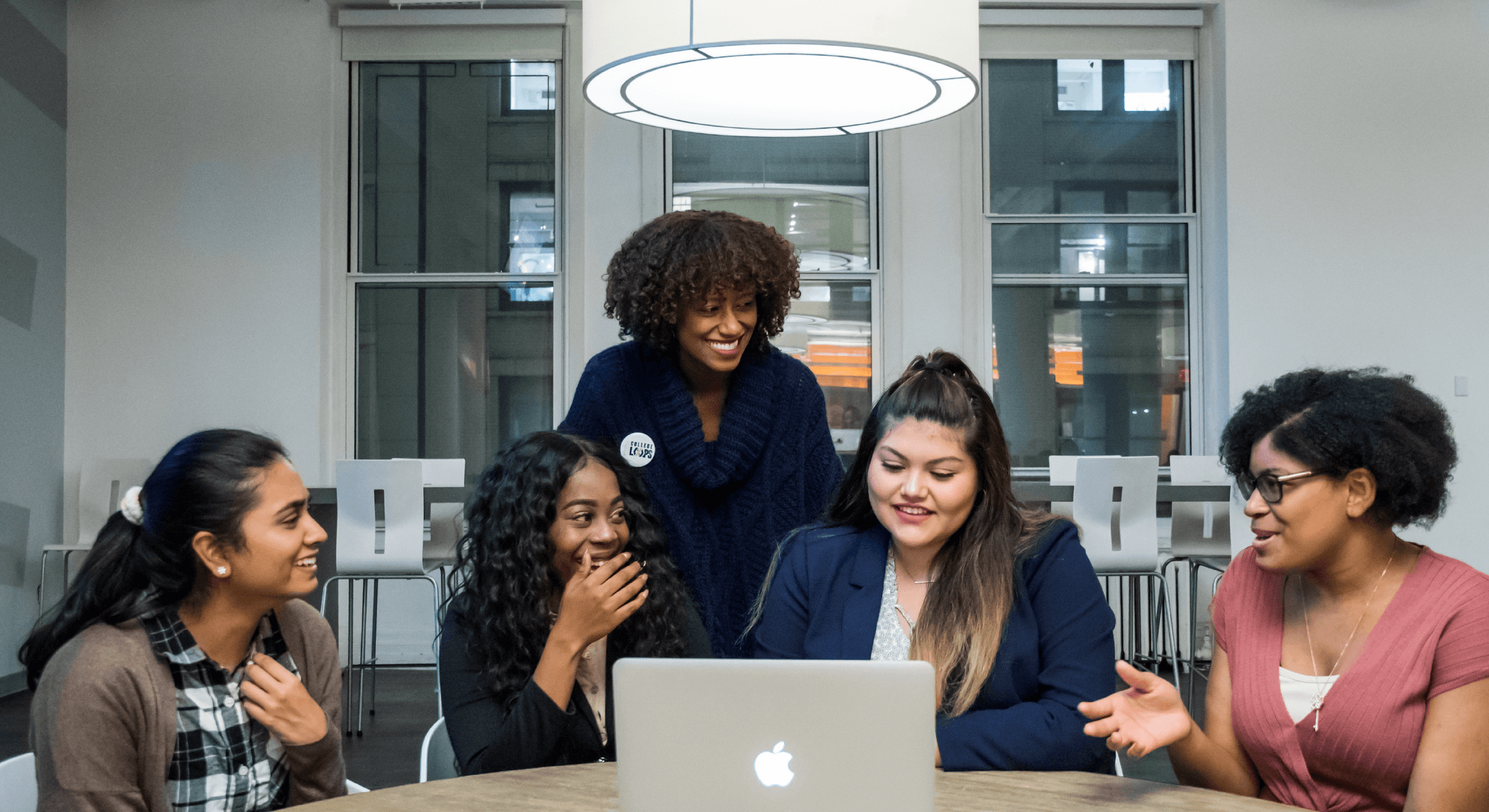 A group of five women, dressed in casual and business attire, gather around a laptop in a modern office space. They are smiling and appear to be engaged in a lively discussion or presentation. A large, round light fixture hangs above their table.