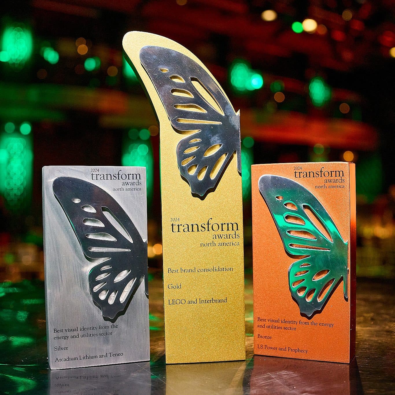 Three awards from the Transform Awards North America are displayed. They feature butterfly designs in gold, silver, and bronze, each labeled with different categories and winners. The background is a dimly lit venue with colorful lights.
