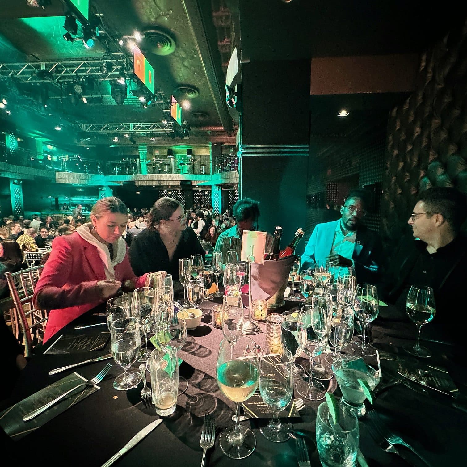 People are seated at a round table in a dimly lit, upscale event space. The table is set with glasses and plates, and there is a bustling crowd in the background. The atmosphere is lively and festive.