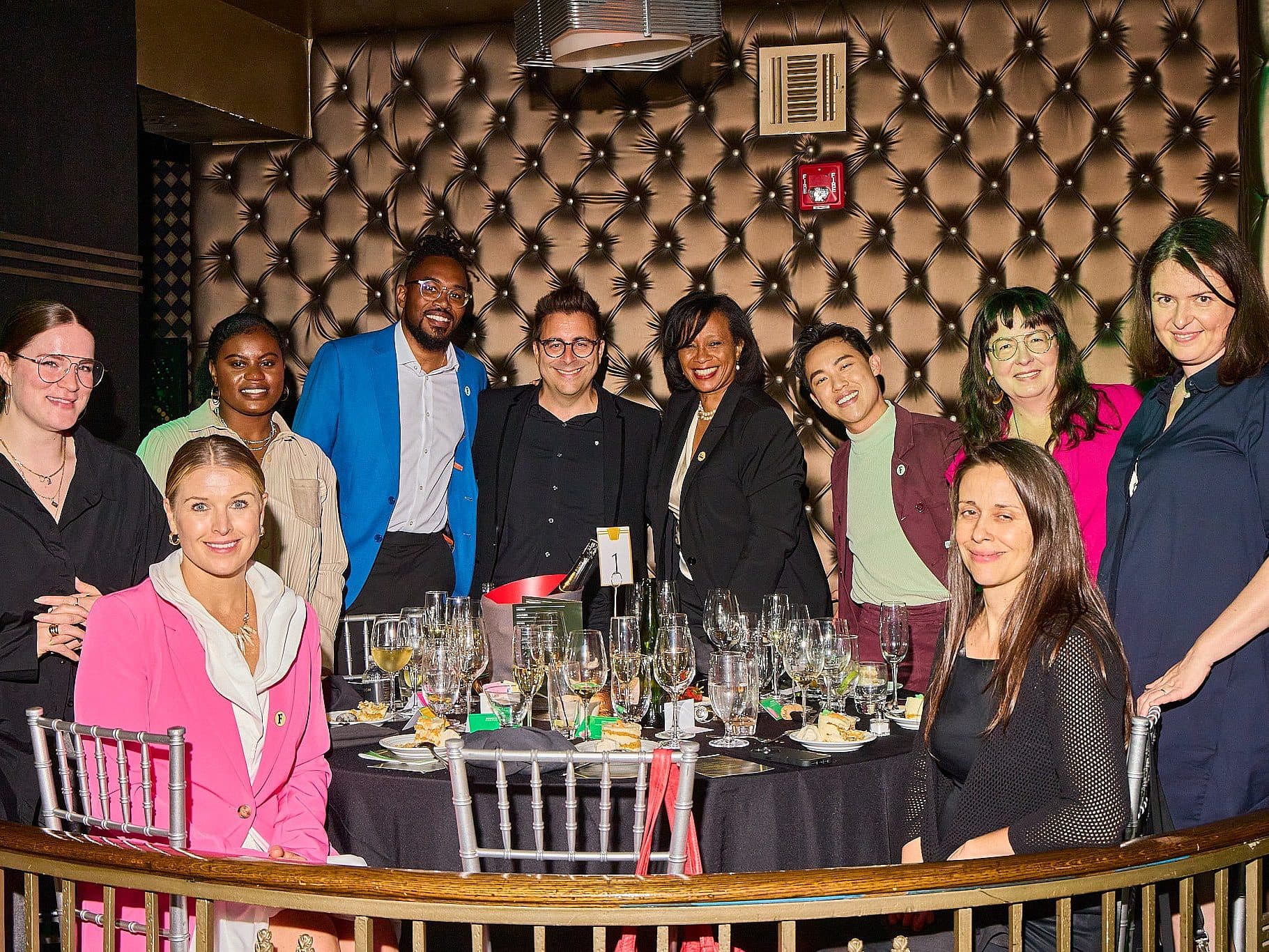 A group of people posing around a table at an event, all dressed in formal attire. The table is set with glasses and plates. The background features a patterned wall. Everyone is smiling, creating a cheerful atmosphere.