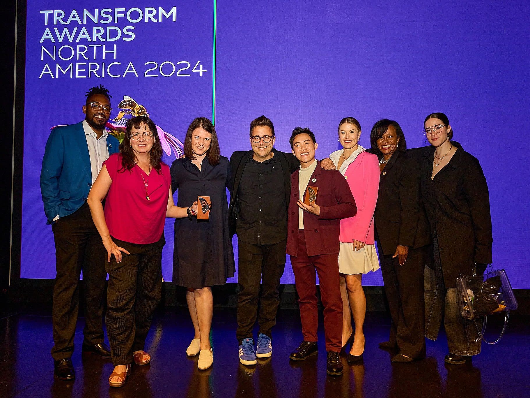 A group of eight people standing together on stage, smiling at the camera. They are dressed in formal attire. Behind them is a blue screen with the text "Transform Awards North America 2024.