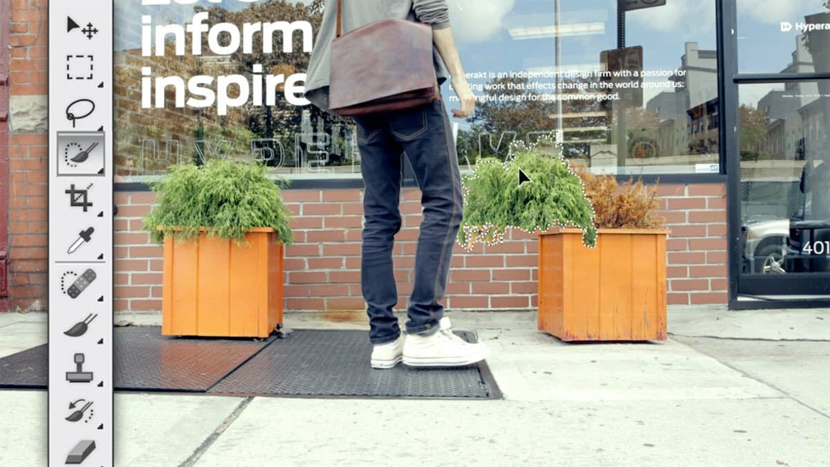 A person wearing jeans and white sneakers stands in front of a brick building with potted plants. A large brown shoulder bag hangs from their shoulder. The building has large windows with text that reads, "Inspire. Inform. Inspire." A toolbar is visible on the left side.