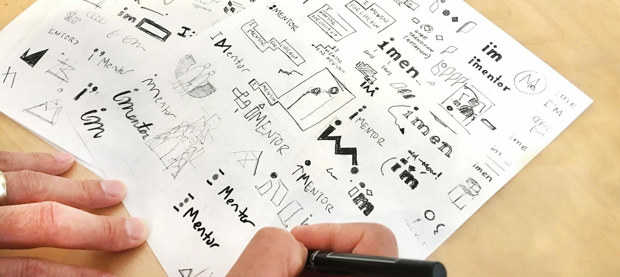 A person's hands are seen sketching logo concepts on multiple sheets of paper. The sketches include various text and icon designs, most of which incorporate the word "iMentor." Different fonts, geometric shapes, and creative illustrations are present on the sketch papers.