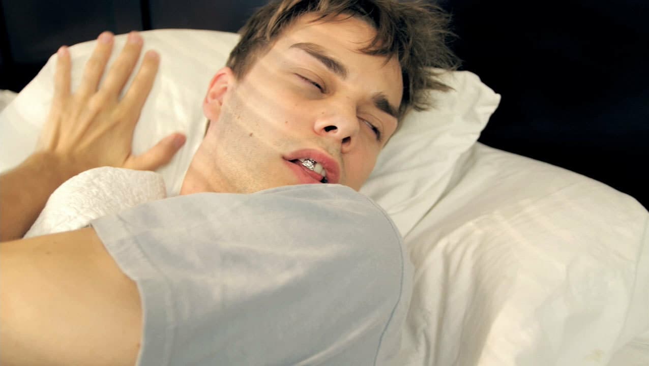 A person lying in bed, appearing to have just woken up. Their eyes are partially open, and sunlight casts shadows across their face. They have messy hair and are resting on a white pillow with white sheets.