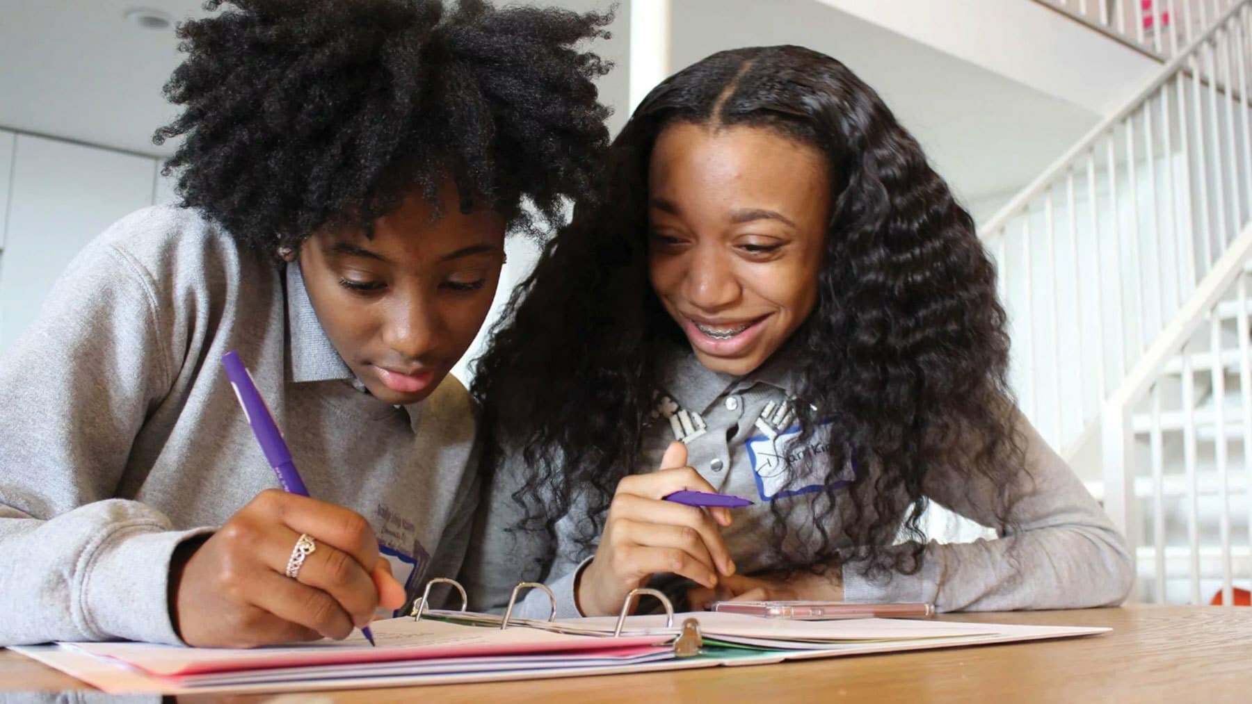 Two students sit at a table, focused on writing in their notebooks. One, with curly hair, writes intently while the other, with long wavy hair, smiles and looks on. Both wear grey sweaters and hold purple pens, with a white background featuring stairs.