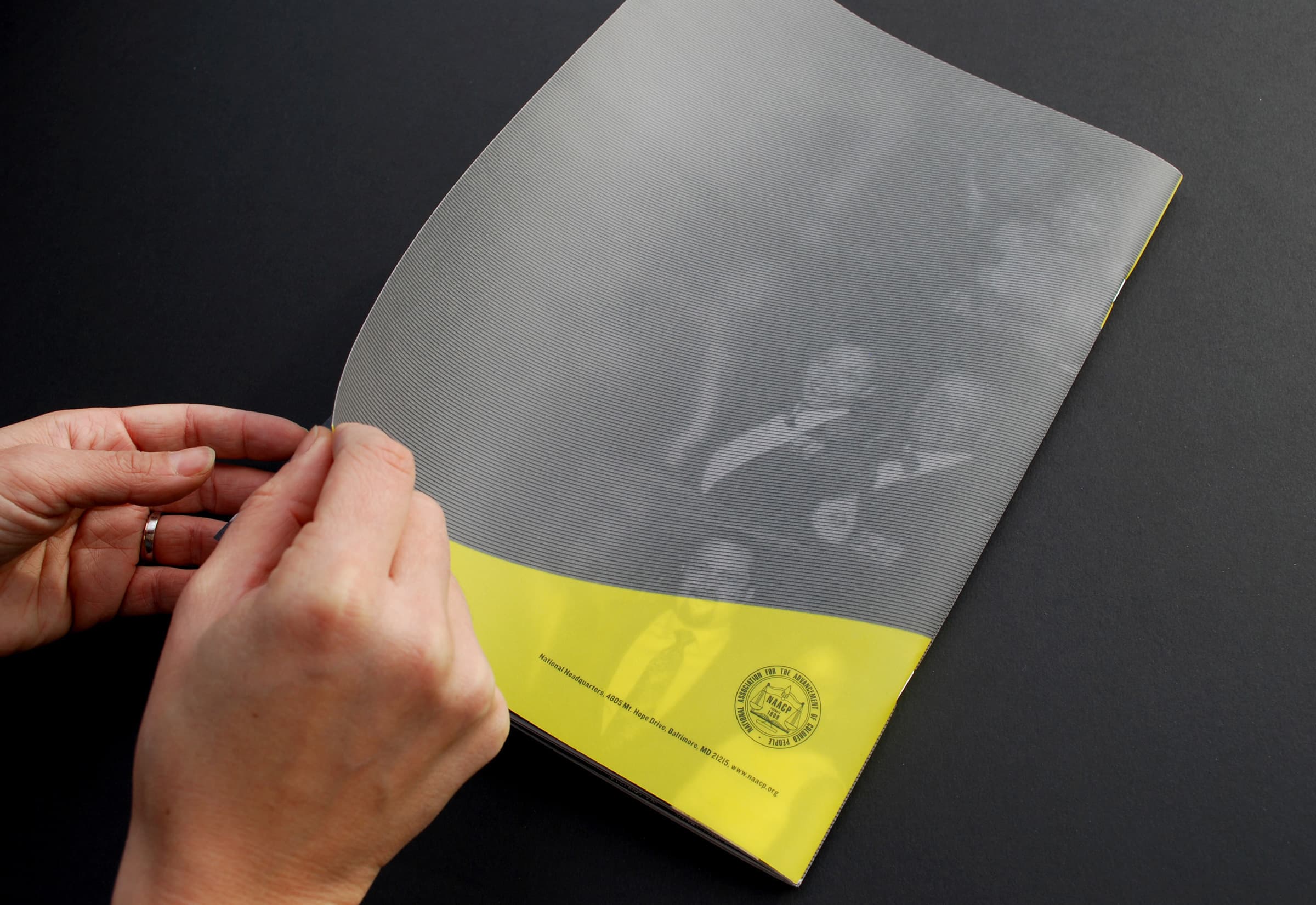 Two hands hold a glossy brochure with a gray and yellow cover. The cover features faint images of people and a circular seal at the bottom with text. The background is black, allowing the brochure to stand out clearly.