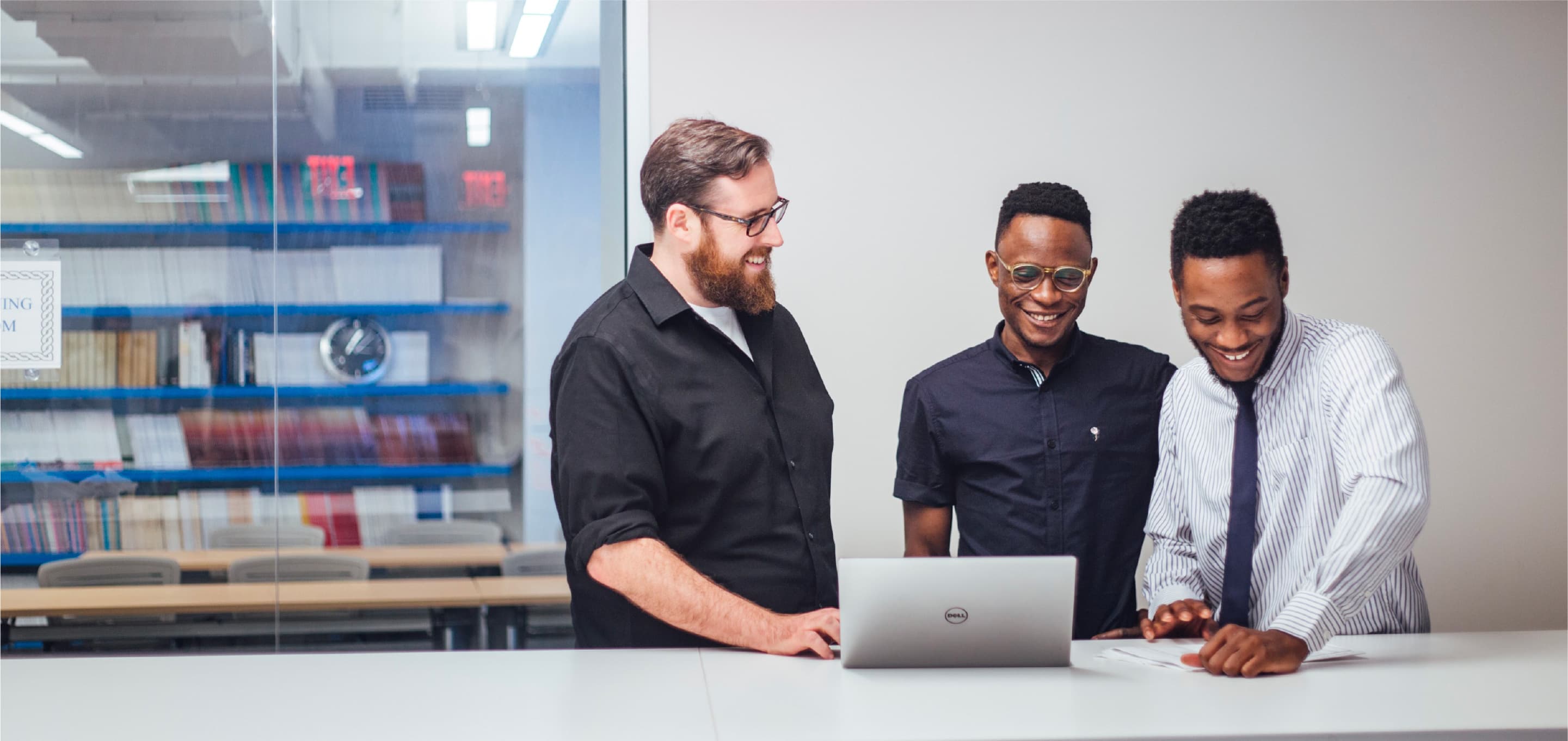 Three men are standing around a table with a laptop, smiling and looking at a document. They are in a modern office setting with a bookshelf in the background. One man wears glasses and a beard, another has glasses, and the third is dressed in a tie and white shirt.