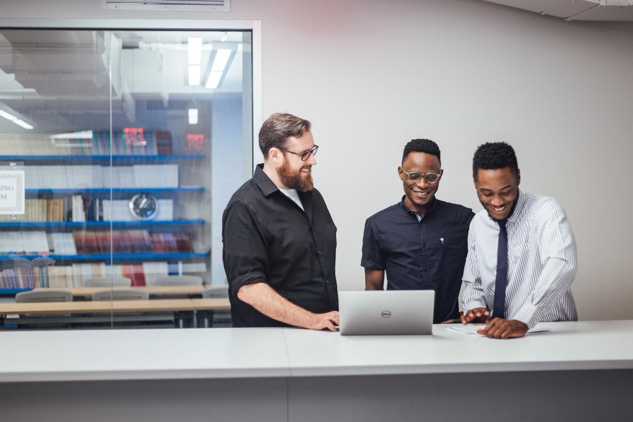 Three men stand around a laptop, engaged in conversation and smiling. They are in an office setting with shelves of books visible through a glass partition in the background. One man wears a black shirt, another a blue shirt, and the third a white dress shirt and tie.