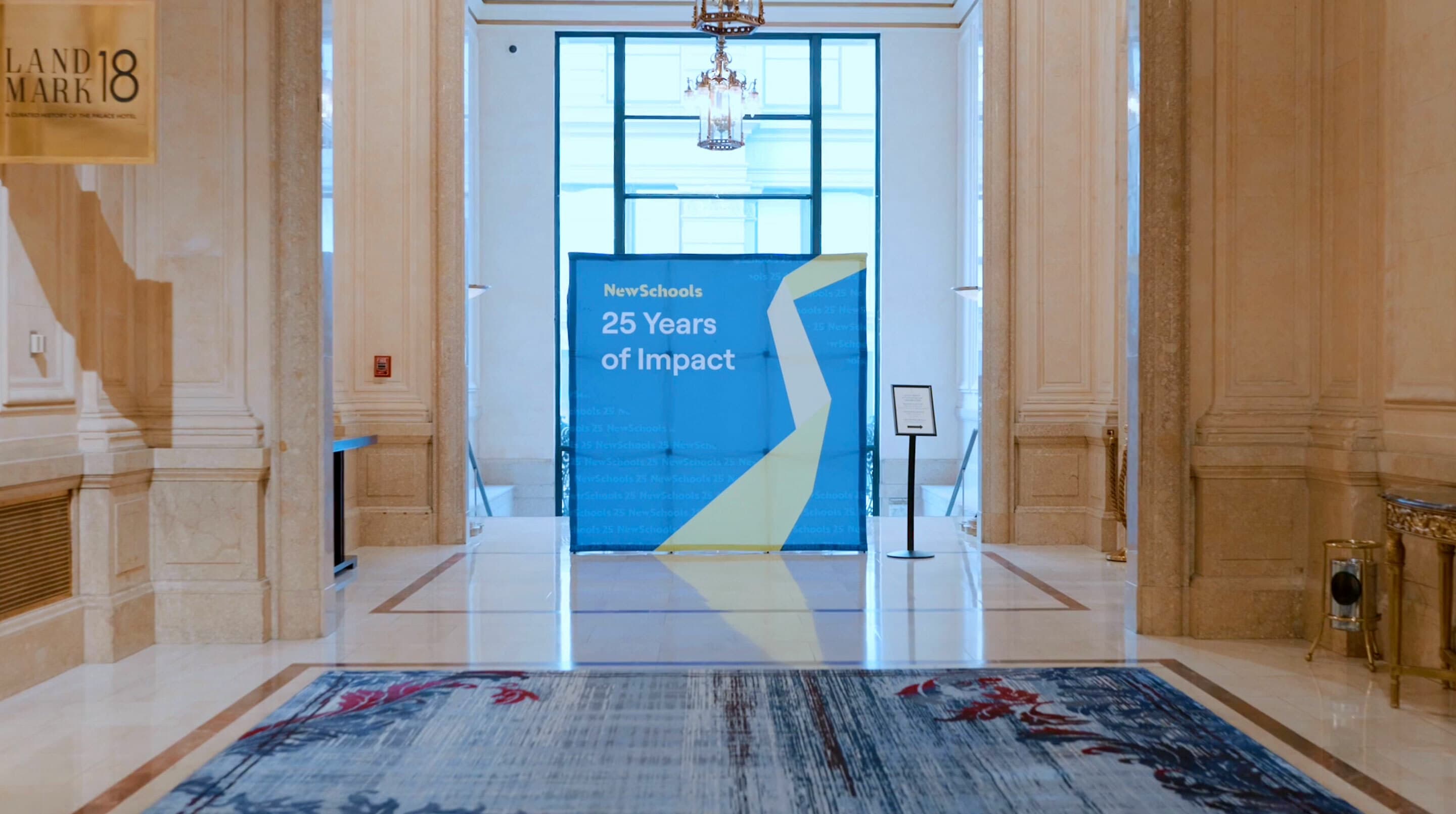 A blue and yellow banner is displayed in a spacious, elegant lobby with high ceilings and marble floors. The banner reads "NewSchools 25 Years of Impact." Large windows and a chandelier are in the background, and "Landmark 18" is seen on a wall to the left.