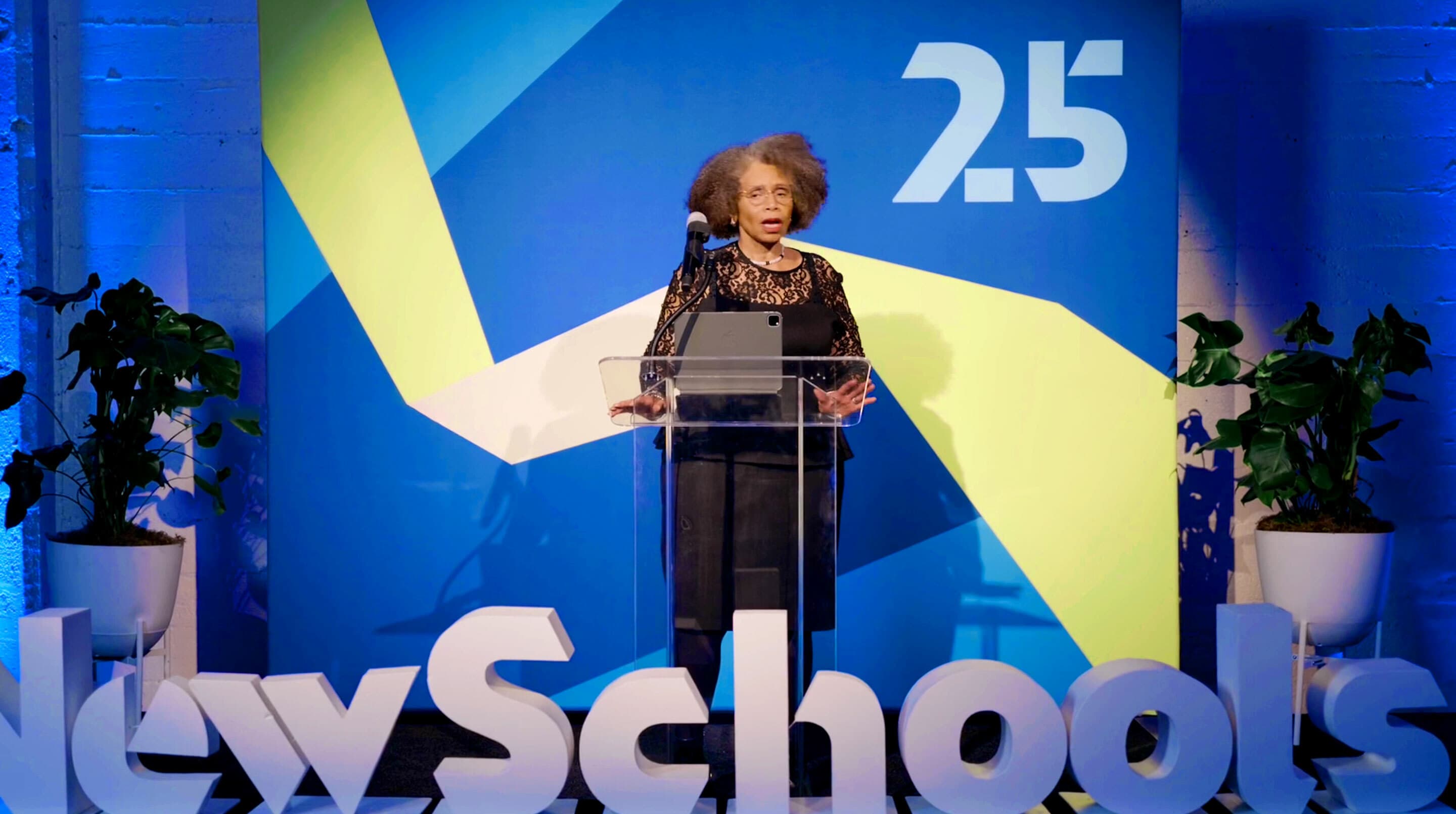 A person with graying hair stands at a clear podium, speaking into a microphone. Behind them is a colorful wall with abstract shapes and the text "2.5." Large letters spelling "New School" are visible in front of the podium.
