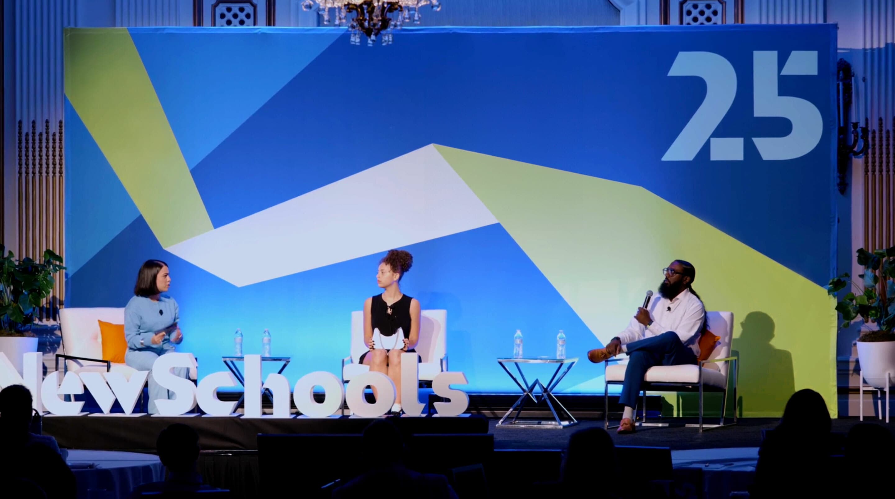 Three people are seated on a stage panel at an event. The blue and green backdrop has the text "NewSchools" and "25." One person speaks into a microphone. Two water bottles are placed on small tables beside them. An audience is visible in the foreground.