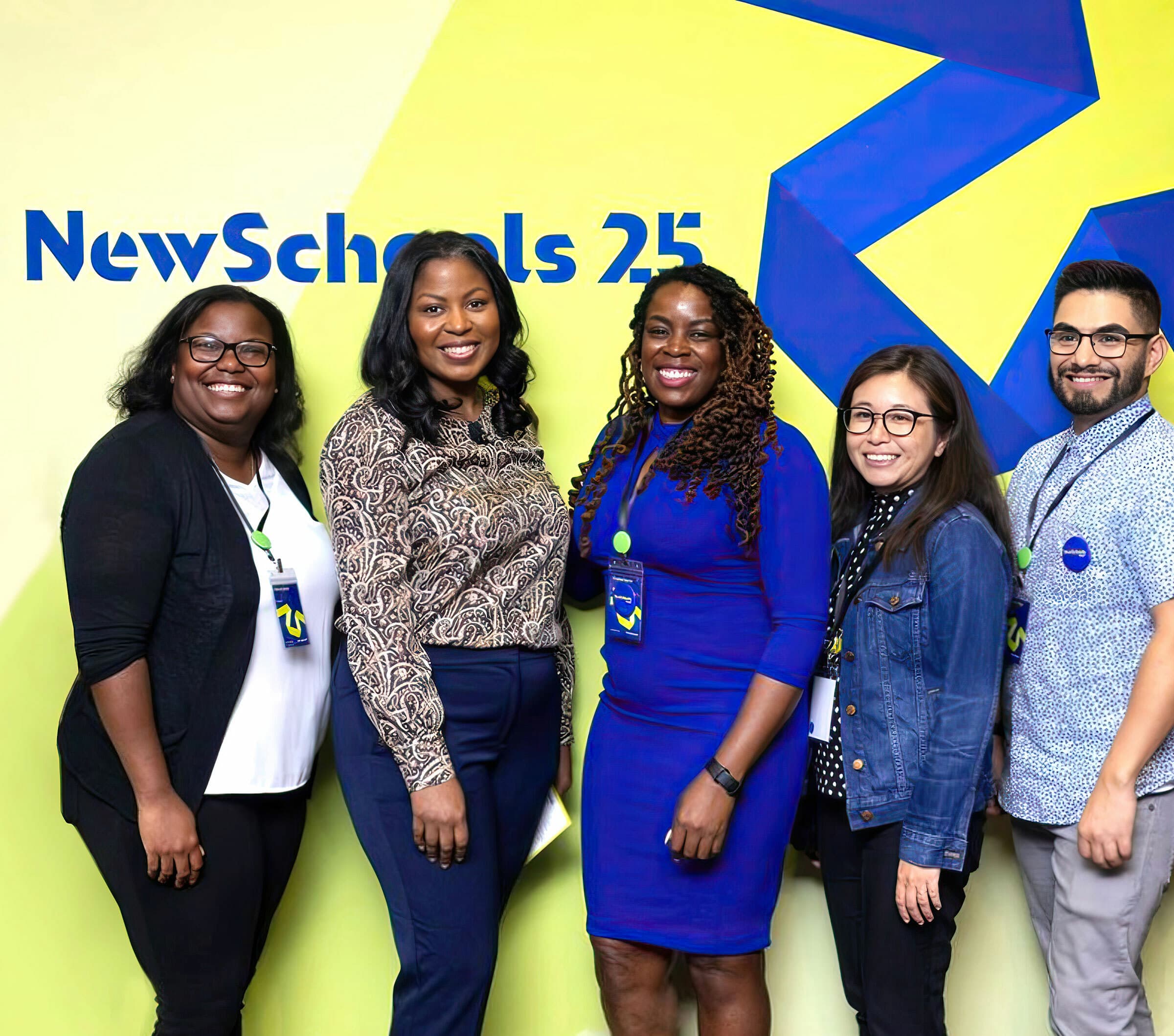 A group of five people stand smiling in front of a colorful wall with the text "NewSchools 25" on it. They are wearing a mix of formal and casual attire, and all have name badges.