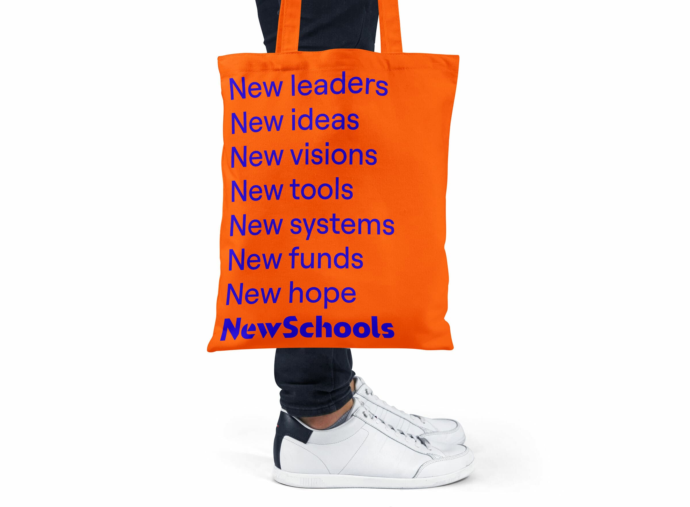 An orange tote bag with blue text reading: "New leaders, New ideas, New visions, New tools, New systems, New funds, New hope. NewSchools." A person in jeans and white sneakers is holding the bag.