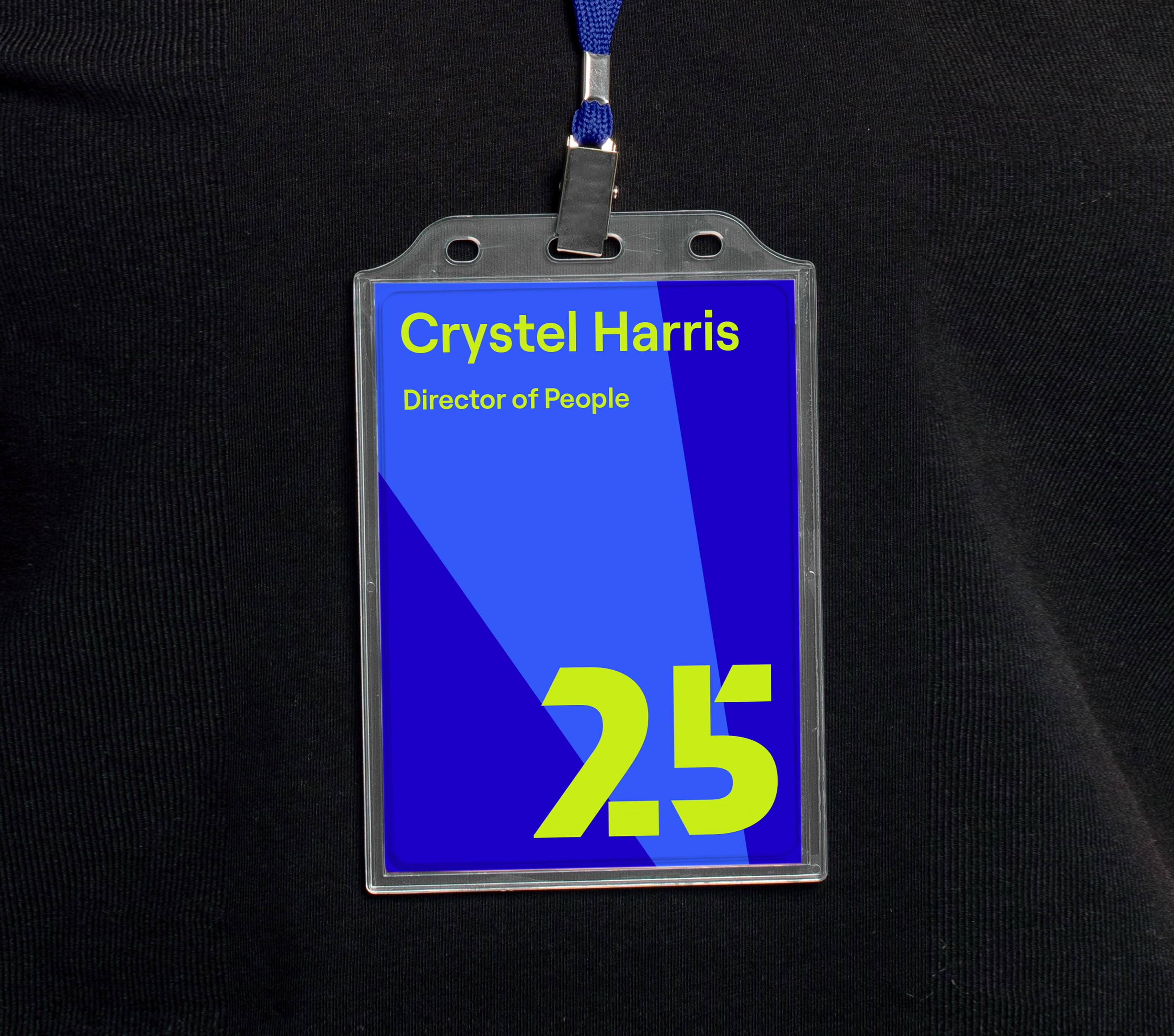 A lanyard with a name badge displays the name "Crystel Harris" and the title "Director of People" against a blue background with green text and abstract shapes, including a large "2.5" in green at the bottom. The badge is worn on a black shirt.