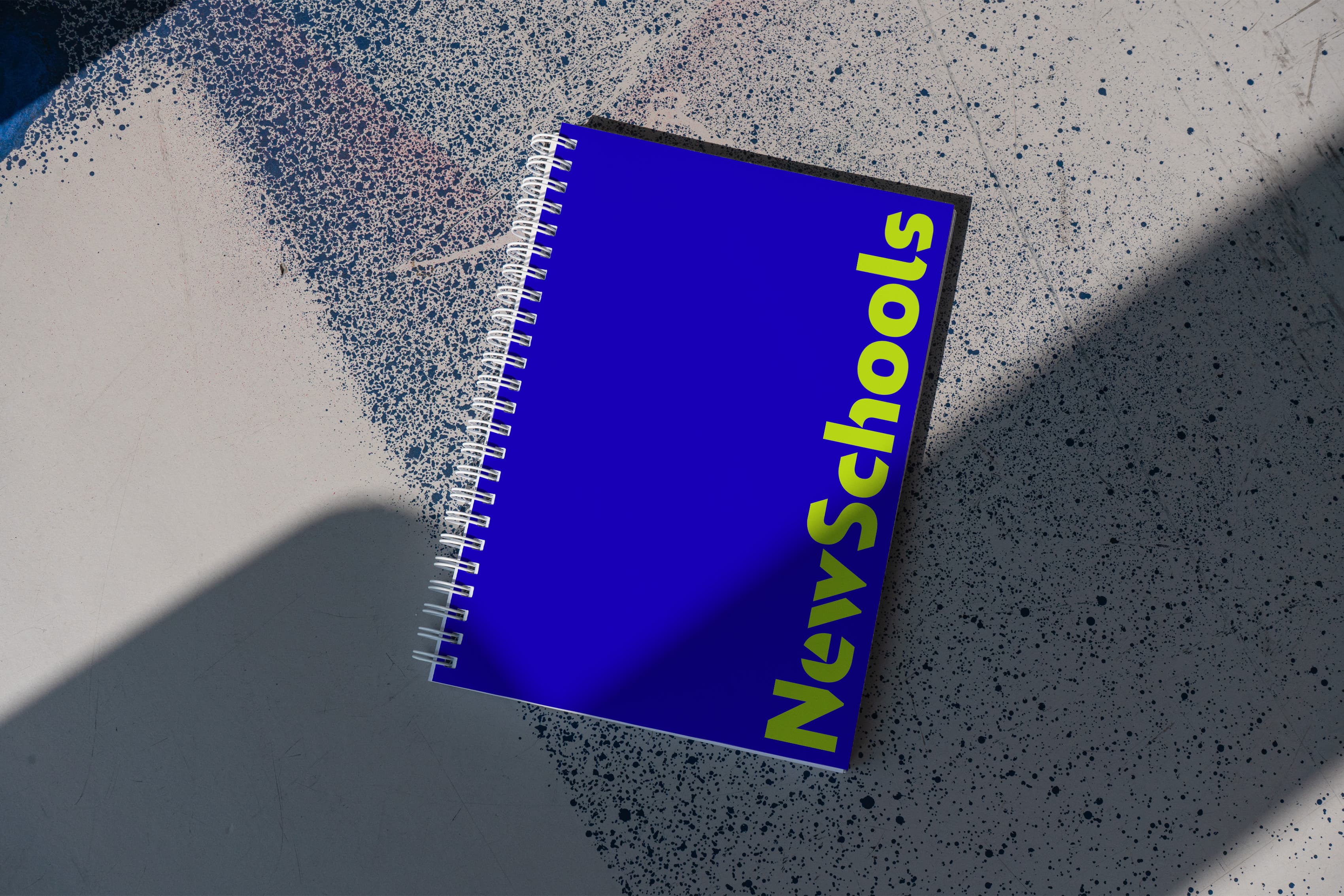 A blue spiral-bound notebook with the word "NewSchools" written vertically in bold yellow letters is placed on a textured surface with shadows and light patterns.