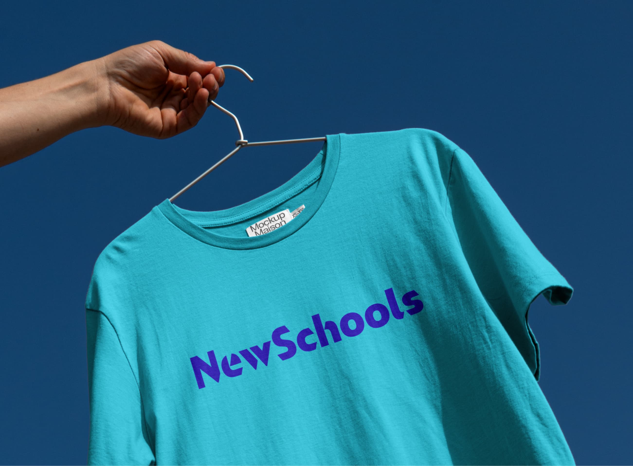 A hand holds a clothes hanger with a turquoise T-shirt, featuring the text "NewSchools" in blue capital letters on the front. The background is a clear blue sky.